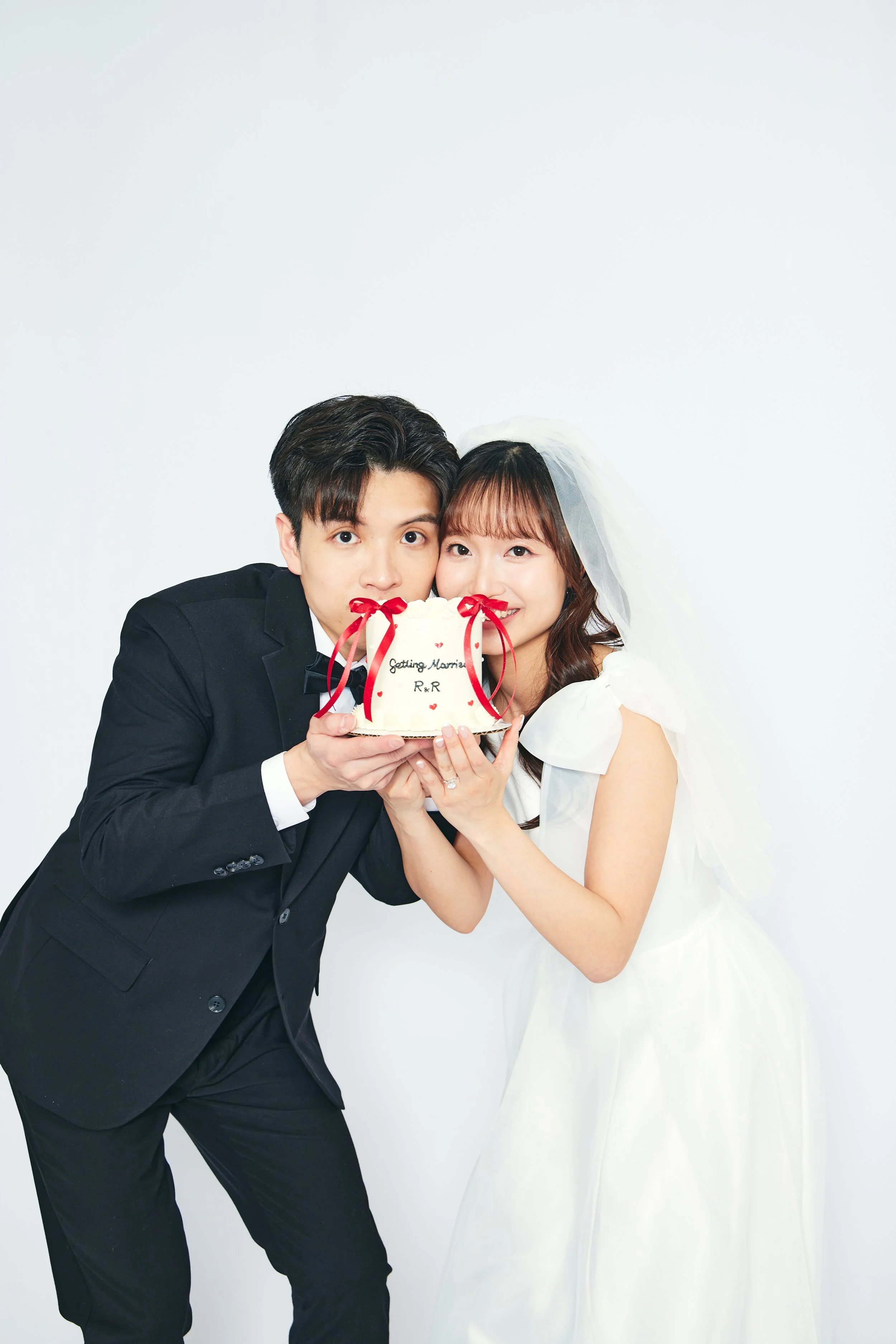 Happy couple in wedding attire holding a wedding cake with red ribbons and writing that says "Getting Married R&R" in front of a white background.