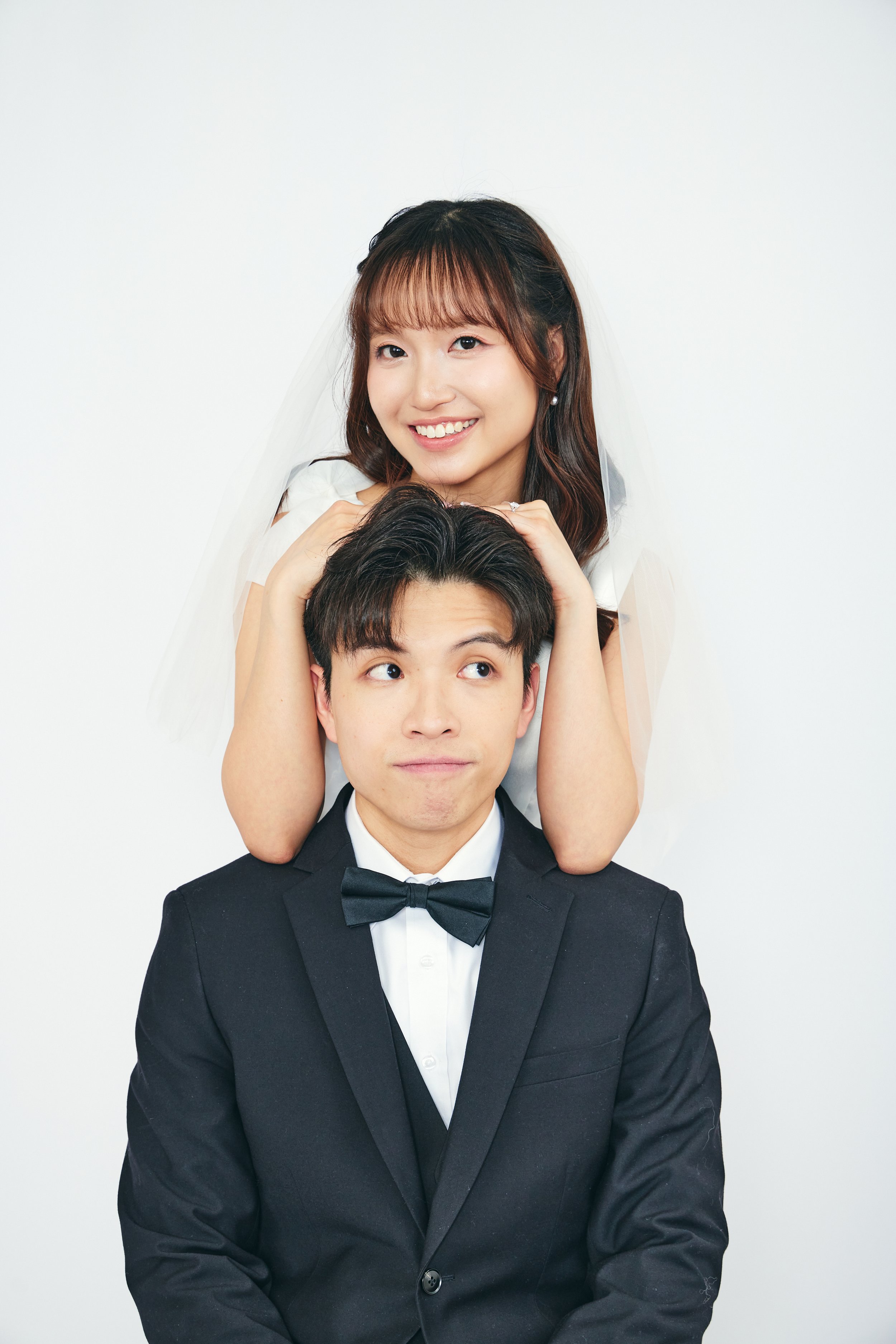 A woman in a wedding dress and veil sitting on a man's shoulders, smiling, with her hands on his head. The man is wearing a black tuxedo with a bow tie and is looking up with a neutral expression against a plain white background.