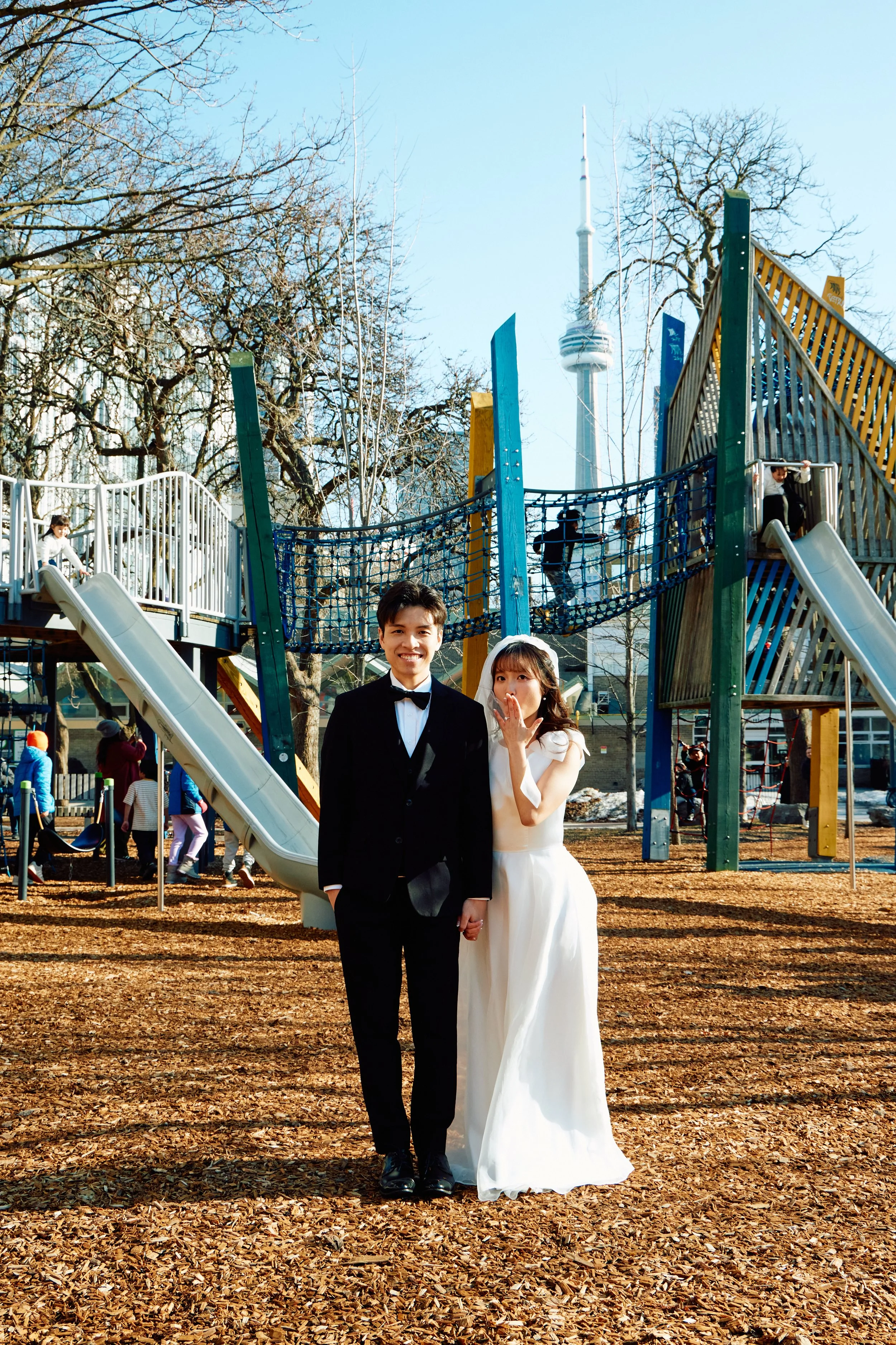 A bride and groom holding hands and standing in a playground with a city skyline and the CN Tower in the background.