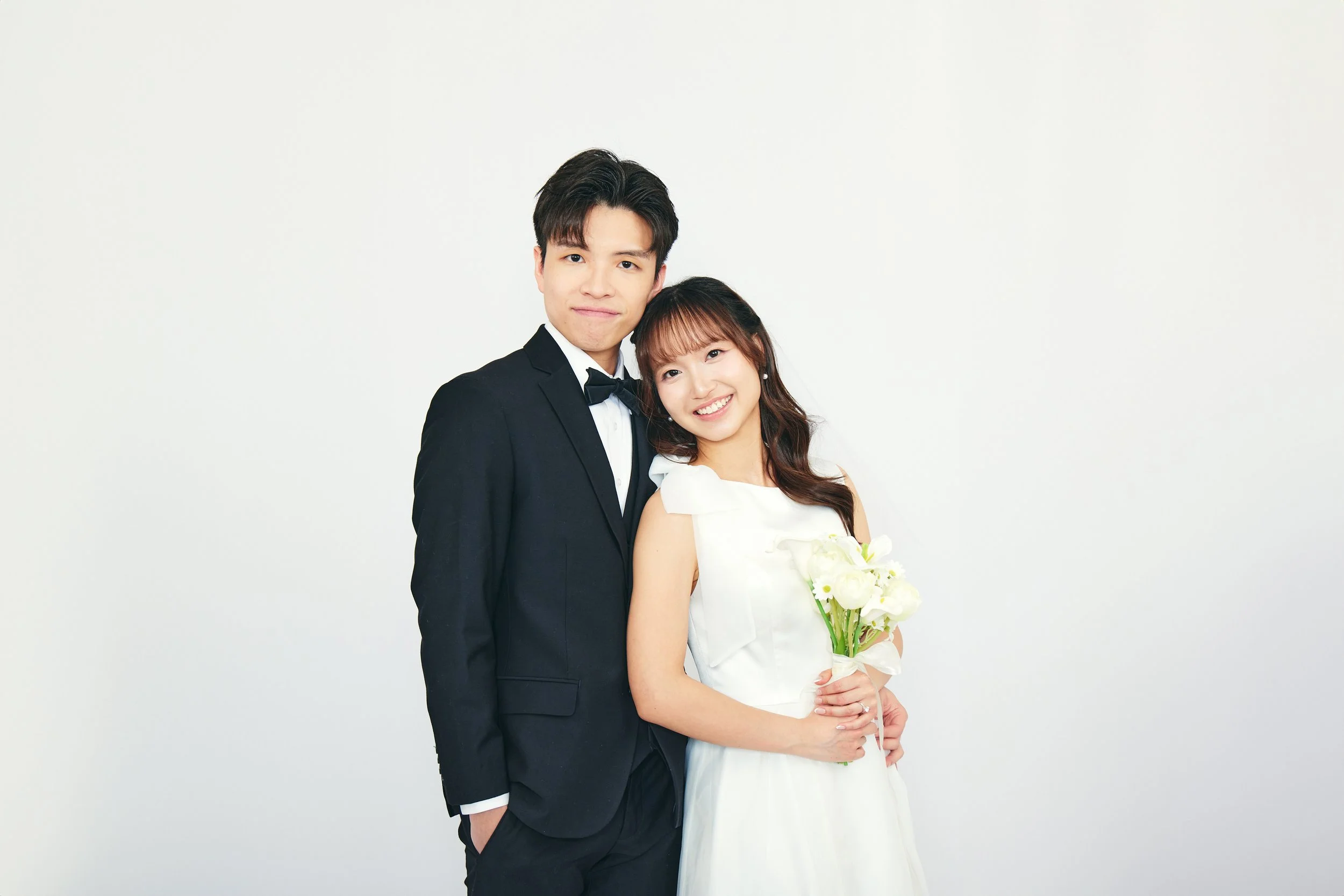 A young man in a black tuxedo with a bow tie standing next to a young woman in a white dress holding a bouquet of white flowers.