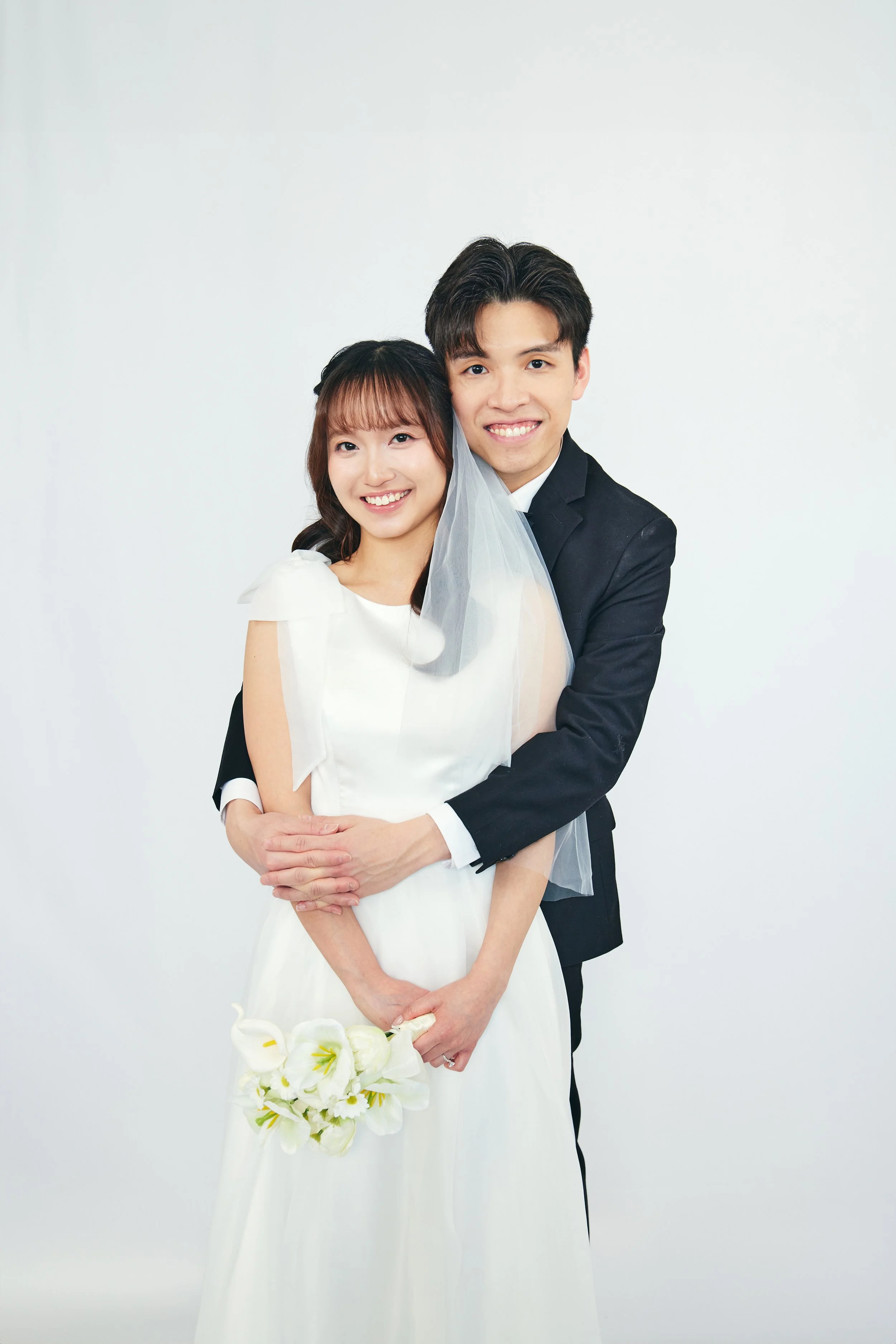 A happy bride and groom posing together, the bride holding a bouquet of white flowers, and the groom wrapping his arms around her, both smiling at the camera.