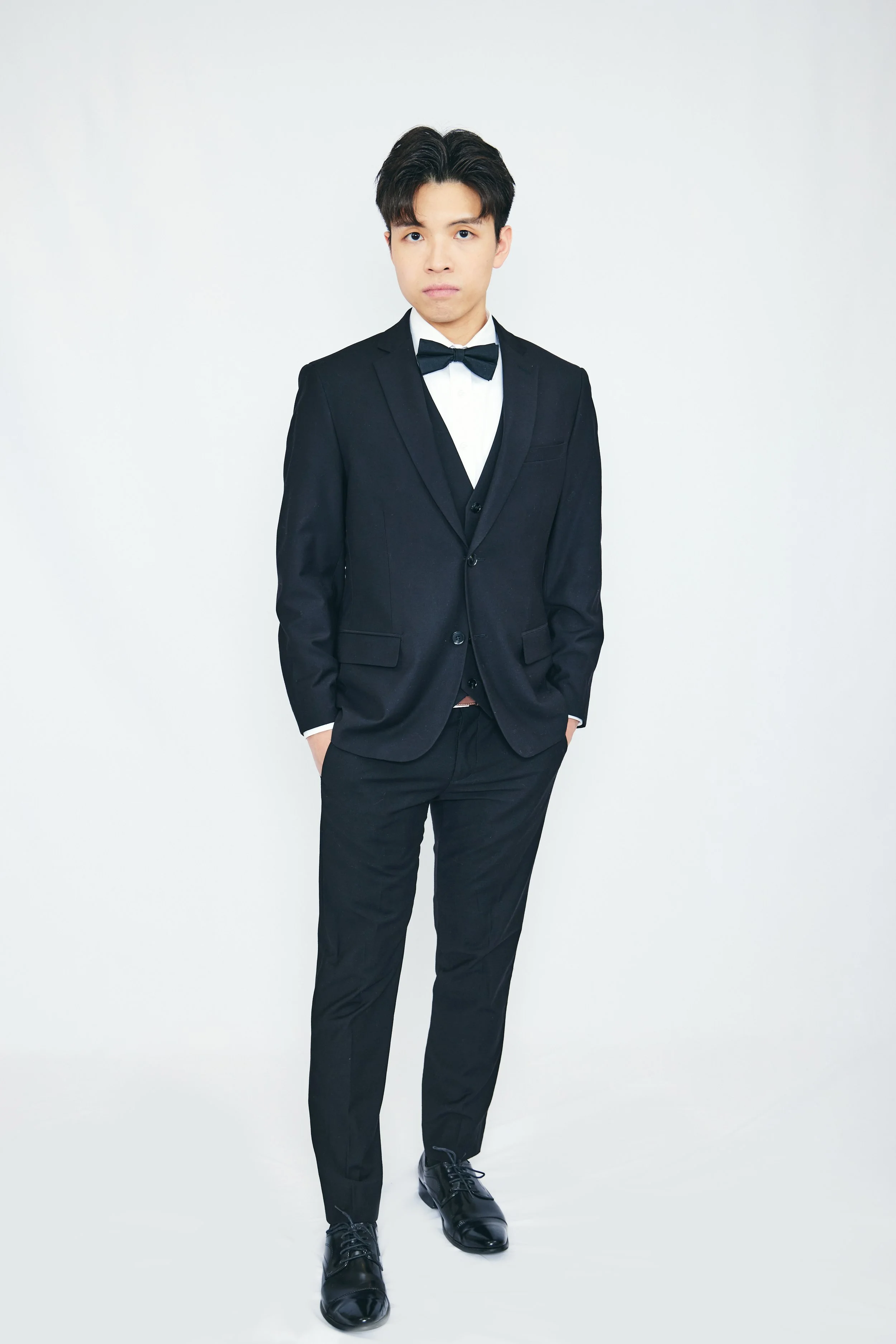 Young man in a black tuxedo with a bow tie, standing against a plain white backdrop.