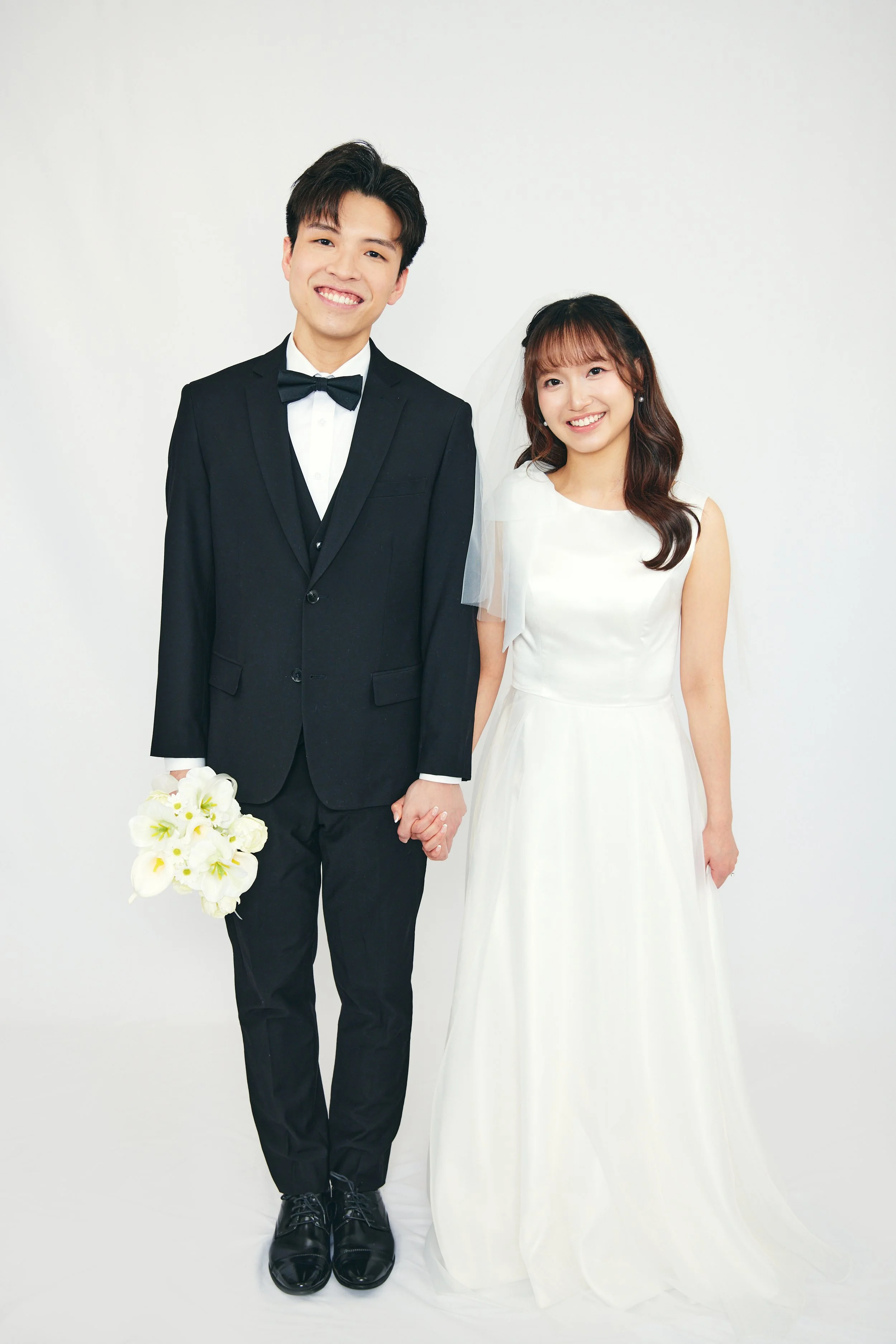 A newlywed couple holding hands, the groom in a black tuxedo with a bow tie, holding a bouquet of white flowers, standing next to the bride in a white wedding gown, smiling against a plain white background.