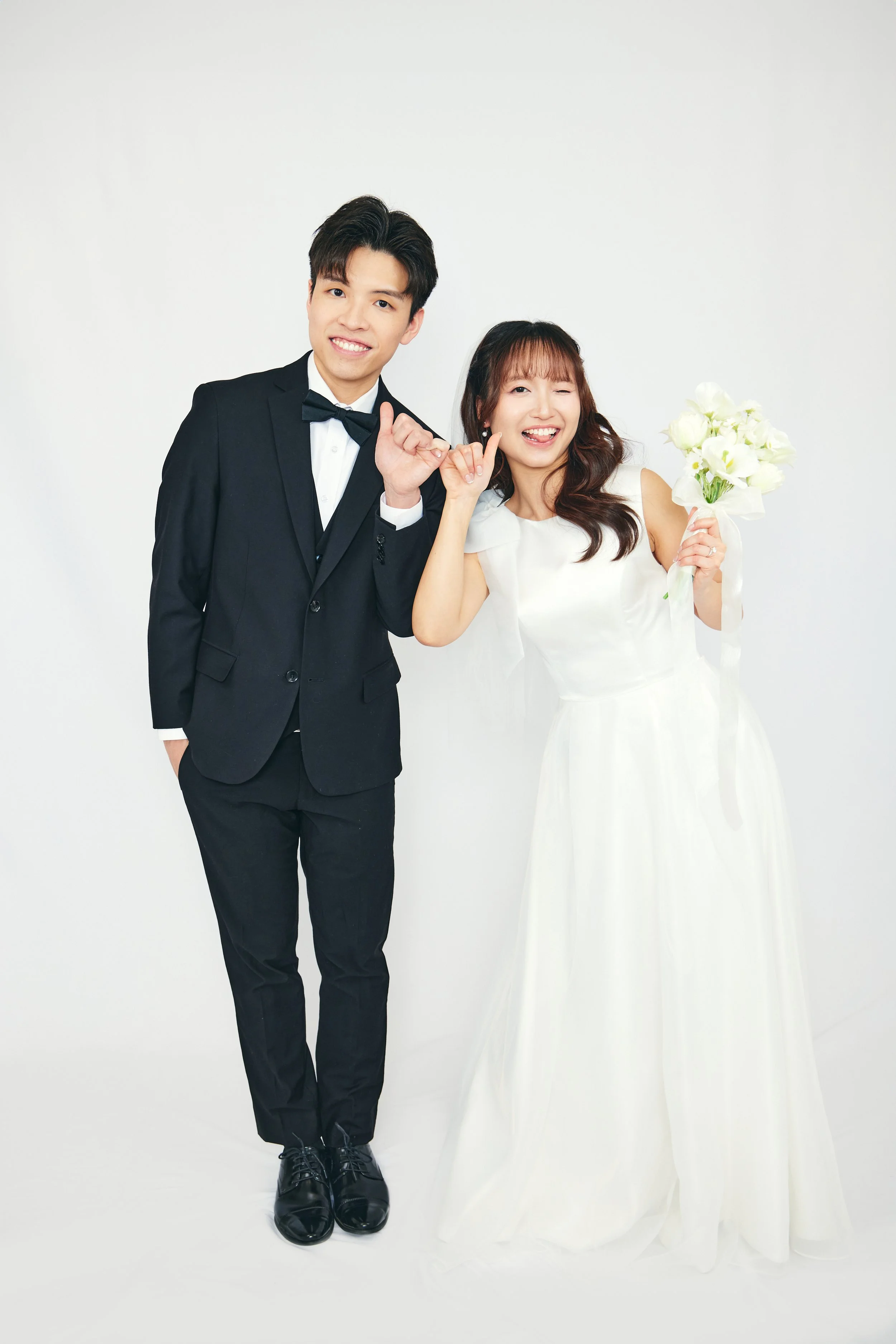 A bride and groom in wedding attire smiling and posing together against a plain white background.