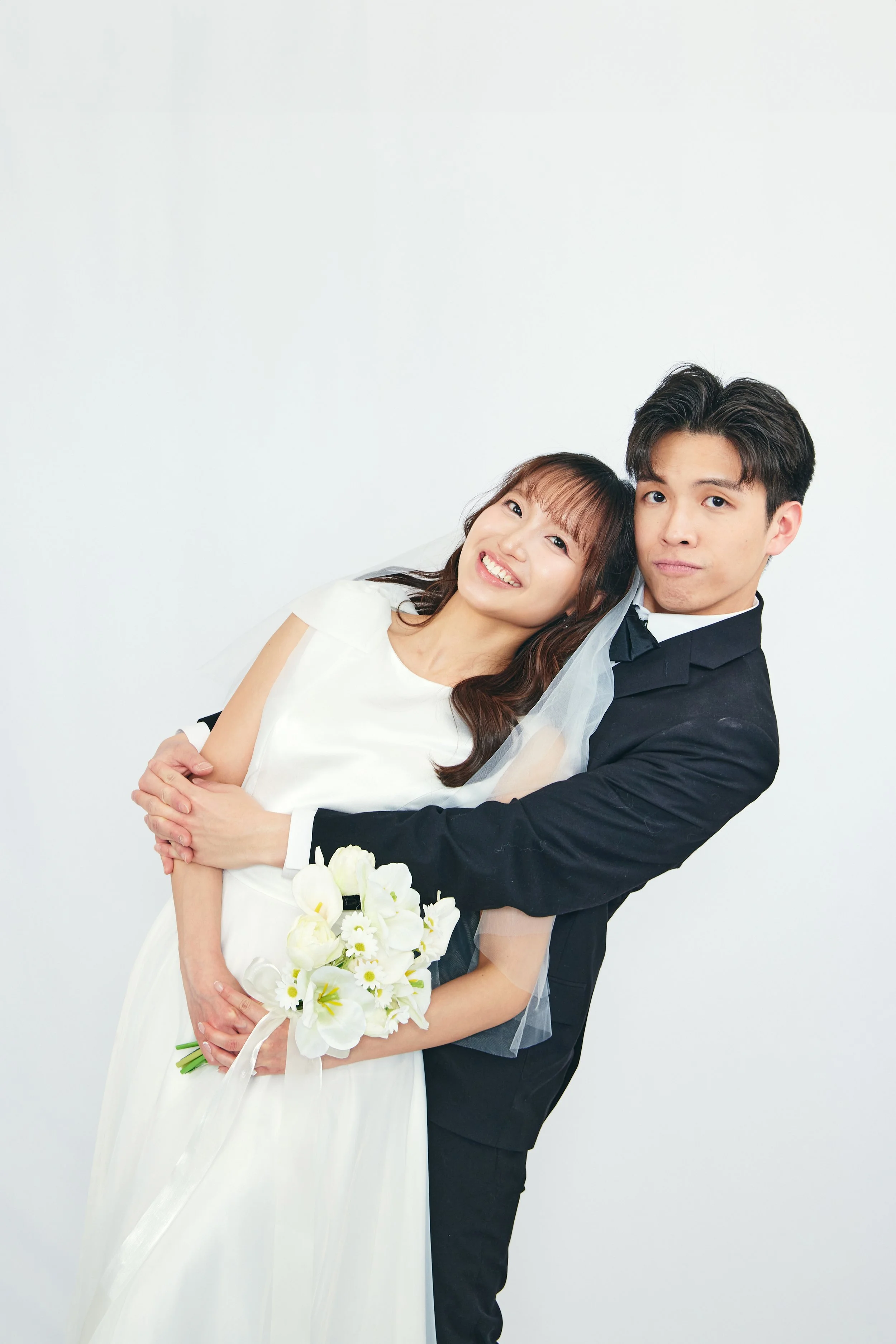 A happy bride in a white dress holding a bouquet of white flowers, being embraced by a groom in a black suit, against a plain white background.
