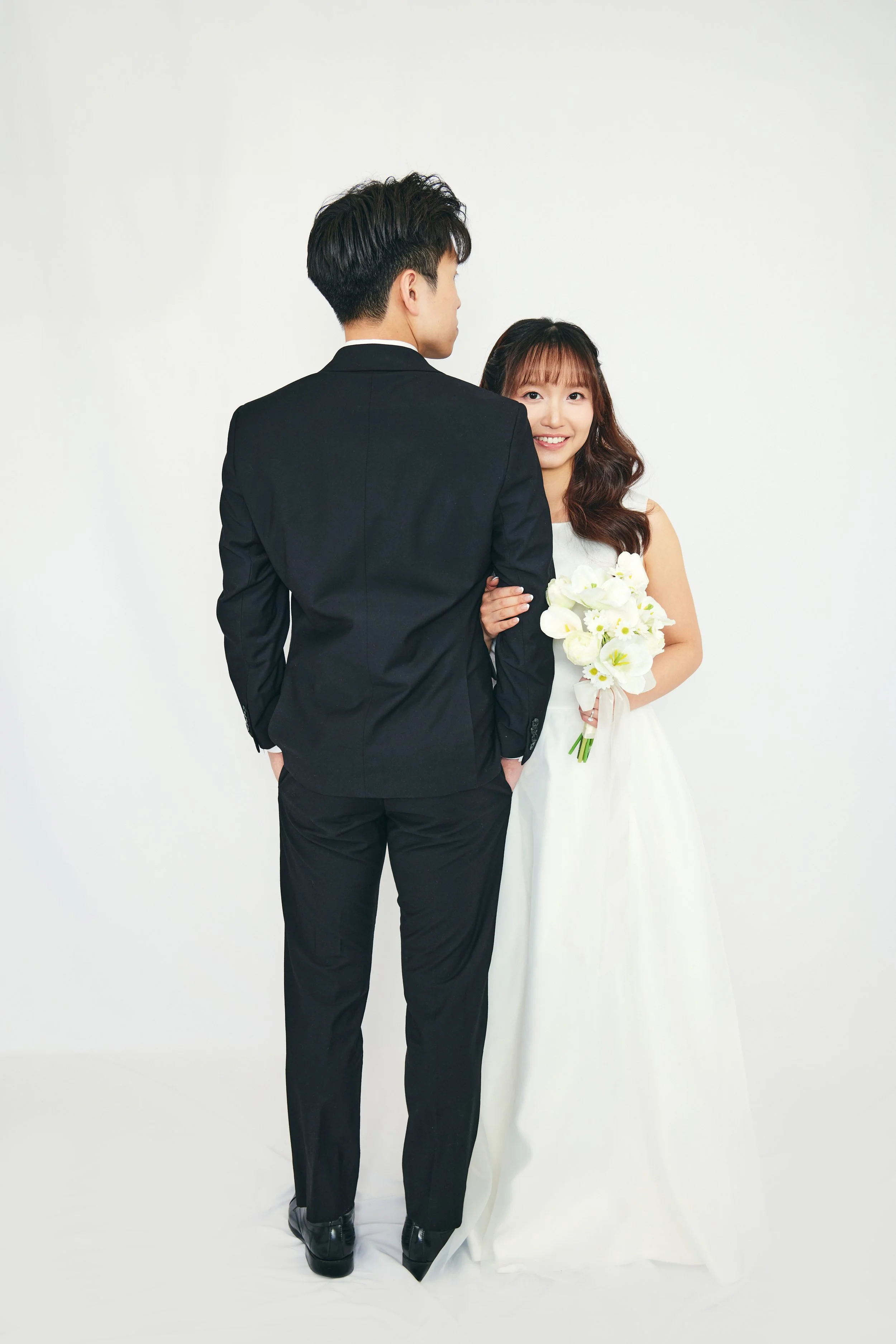 A bride and groom pose for a wedding photo against a plain white background. The bride is holding a bouquet of white flowers and smiling.