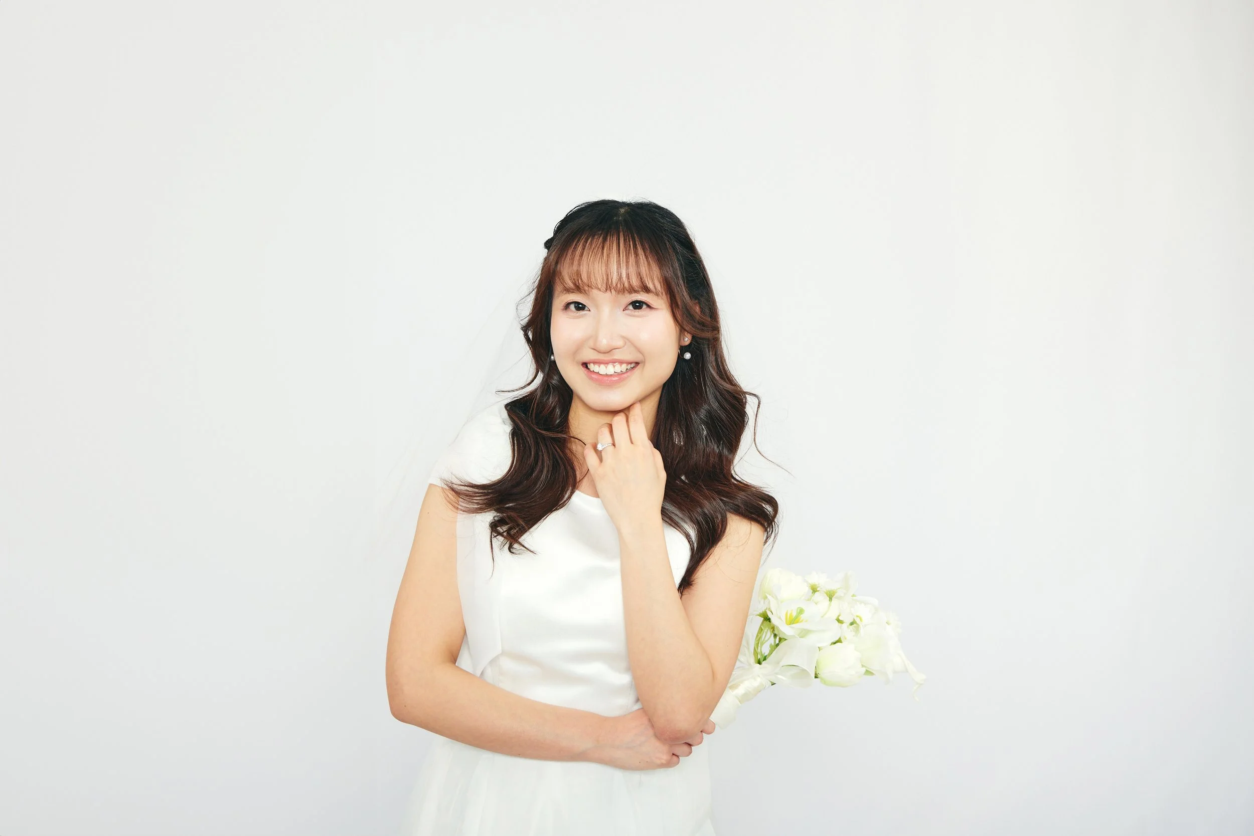 A smiling woman in a white dress holding a bouquet of white flowers against a plain white background.