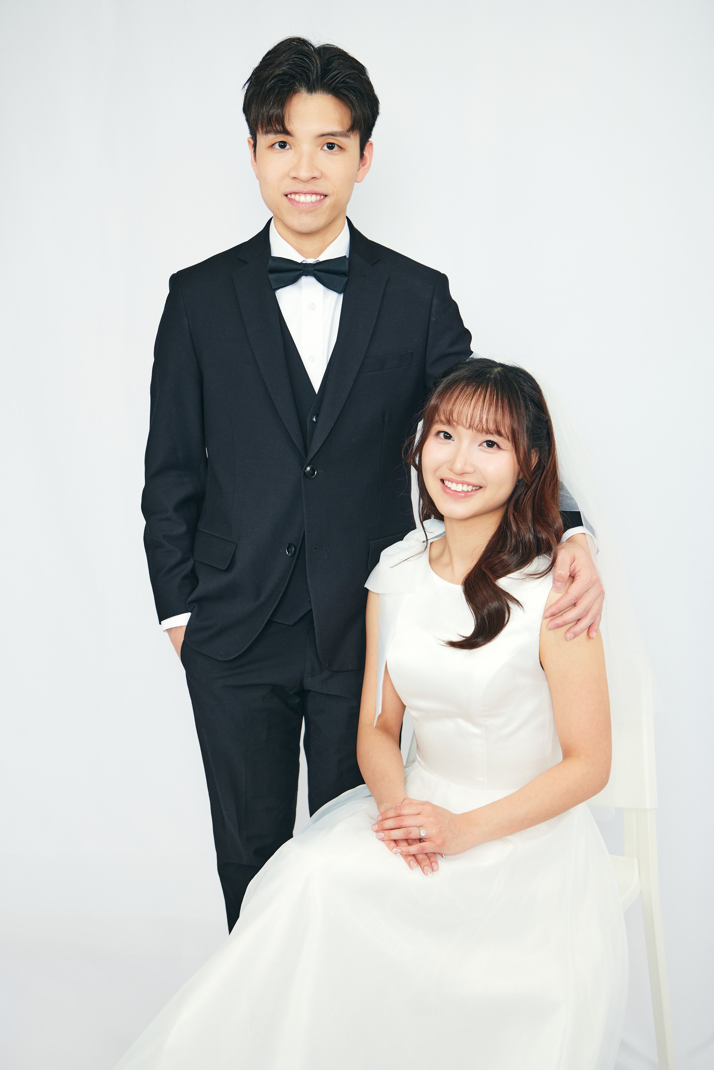 A young man in a black tuxedo with a bow tie stands next to a young woman in a white wedding dress, both smiling at the camera against a plain white background.