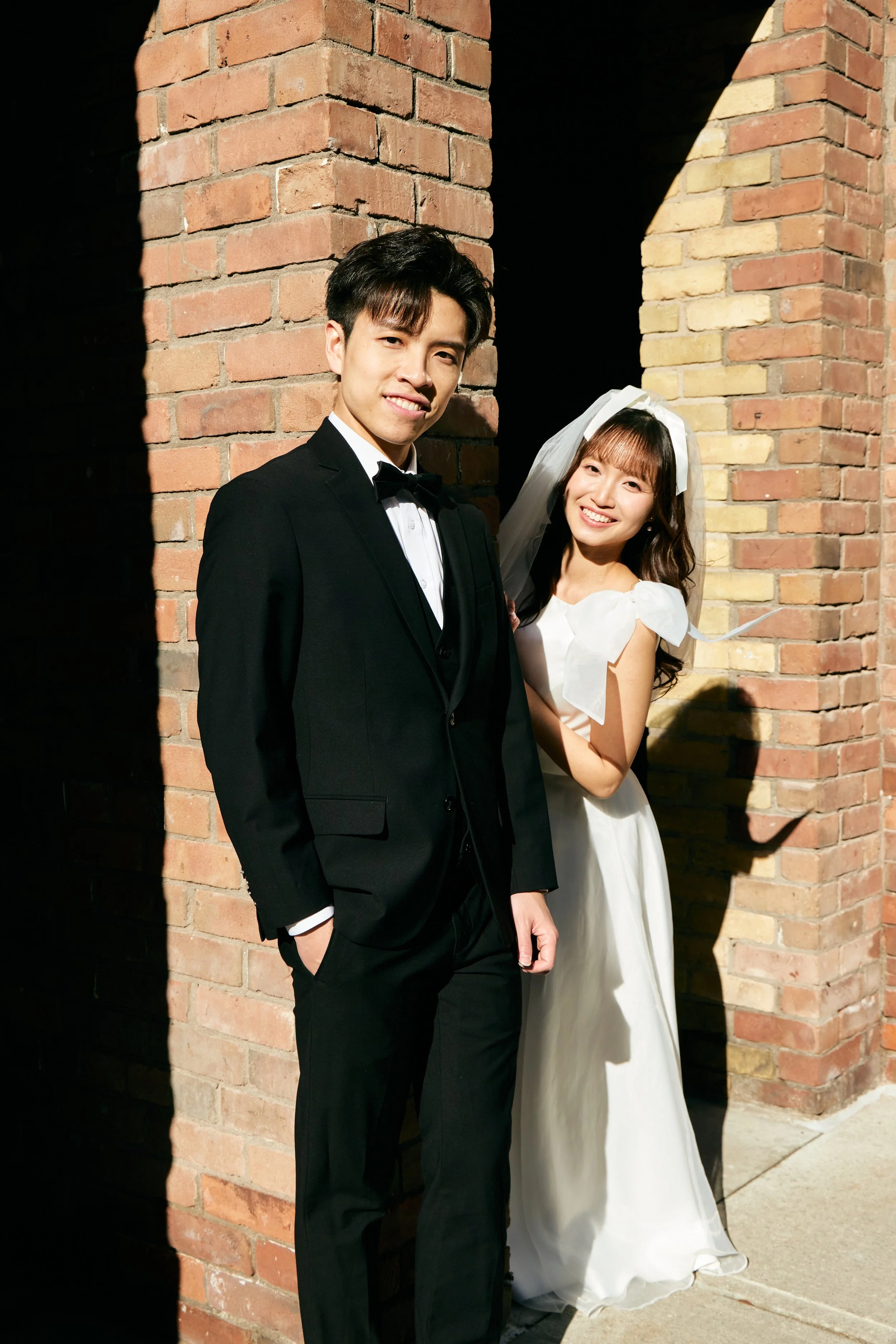 A young man in a black tuxedo with a bow tie and a young woman in a white wedding dress with a veil standing together outside against a brick wall, smiling at the camera.