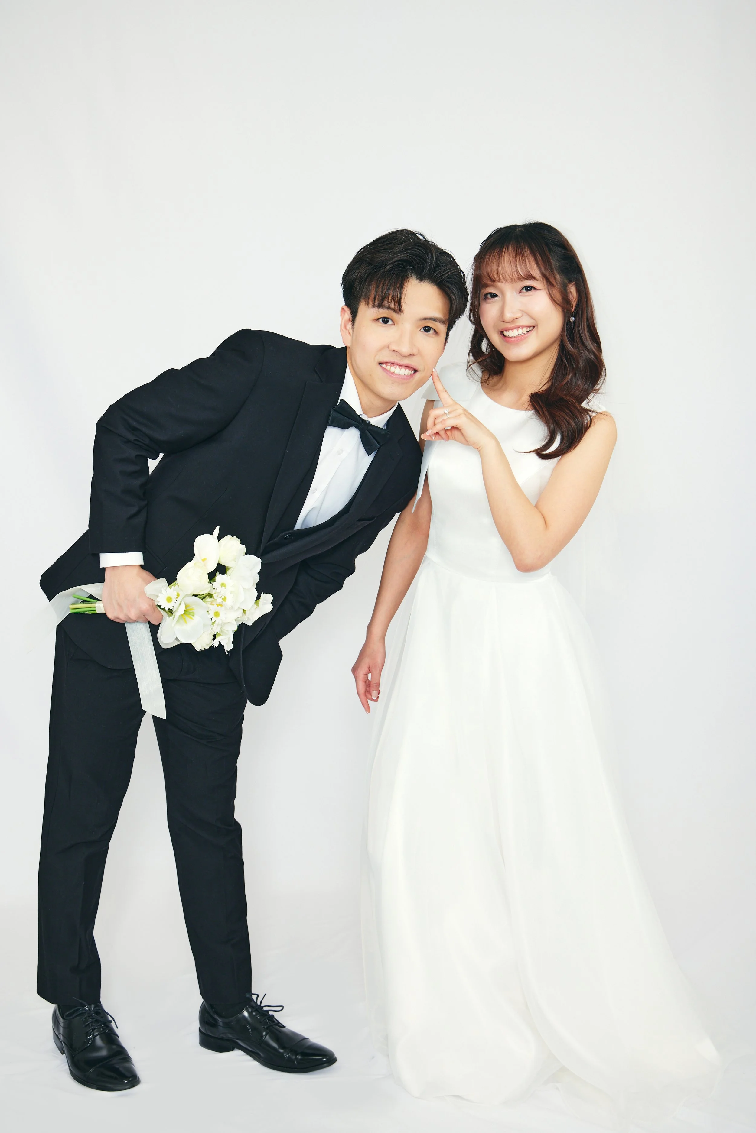 A bride and groom in wedding attire posing together against a plain white background. The groom is wearing a black tuxedo with a bow tie, holding a bouquet of white flowers, and leaning towards the bride. The bride is wearing a sleeveless white weddi