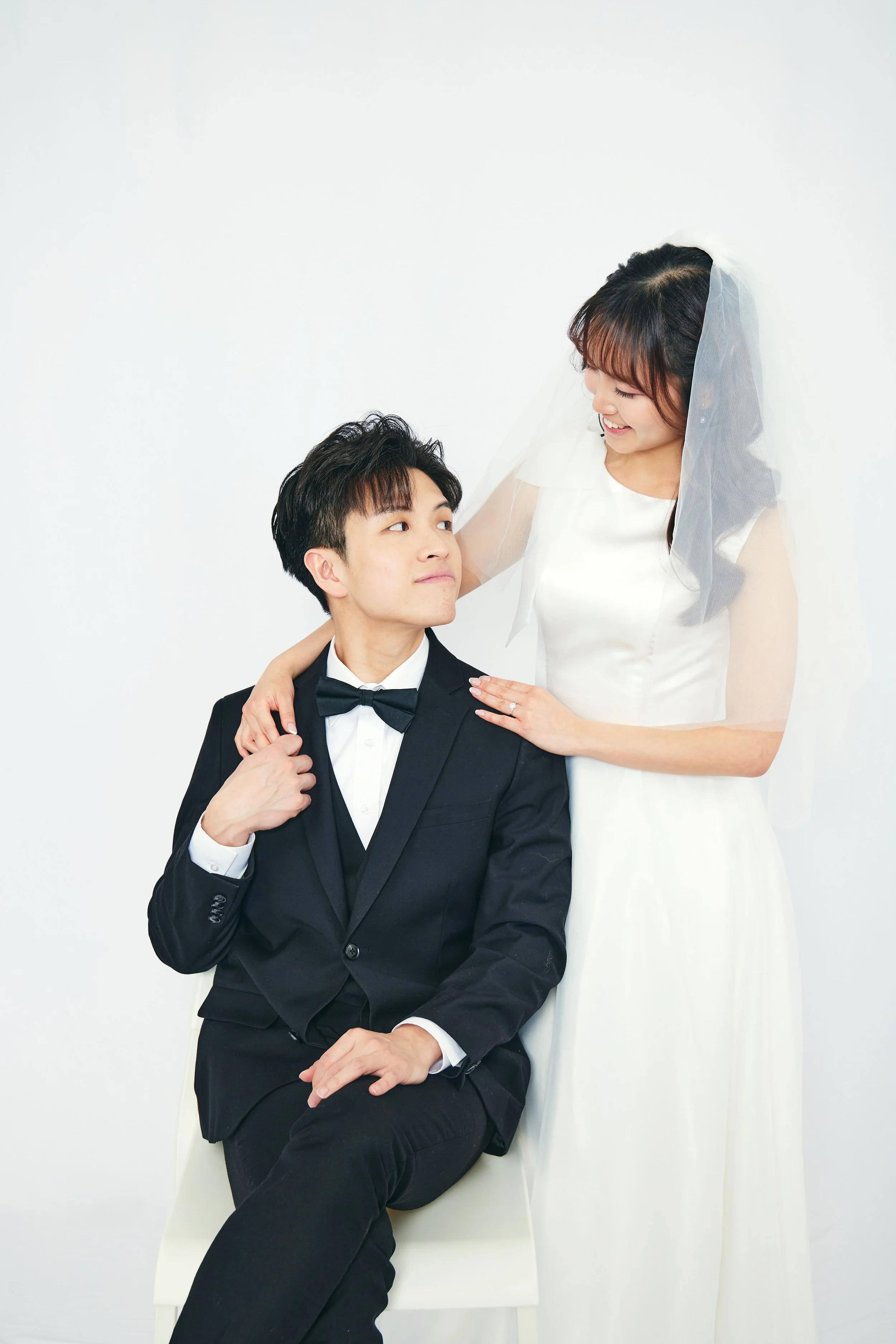 A bride in a white wedding dress with a veil and a groom fitted in a black tuxedo with a bow tie, posing together against a plain white background.