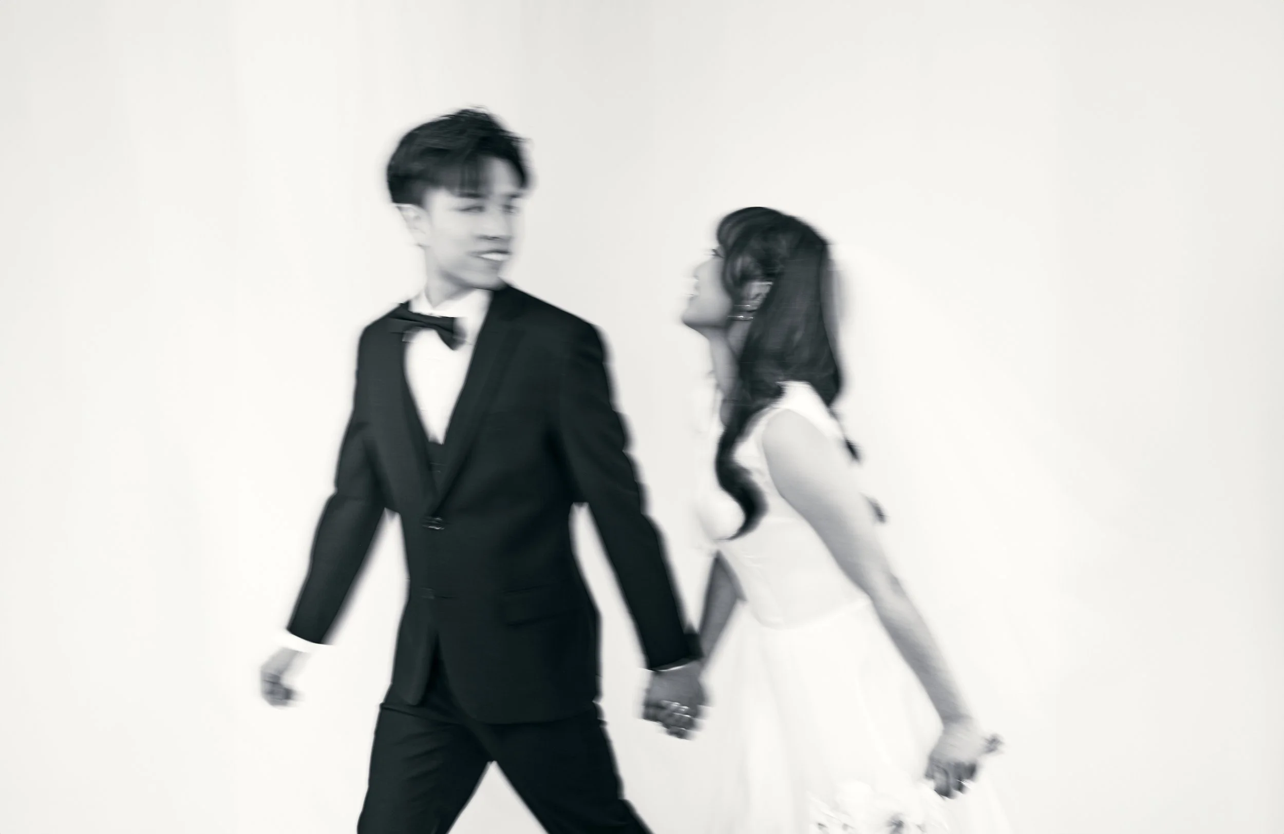 A blurred black and white photo of a man in a tuxedo and a woman in a white dress holding hands and walking together against a plain background.