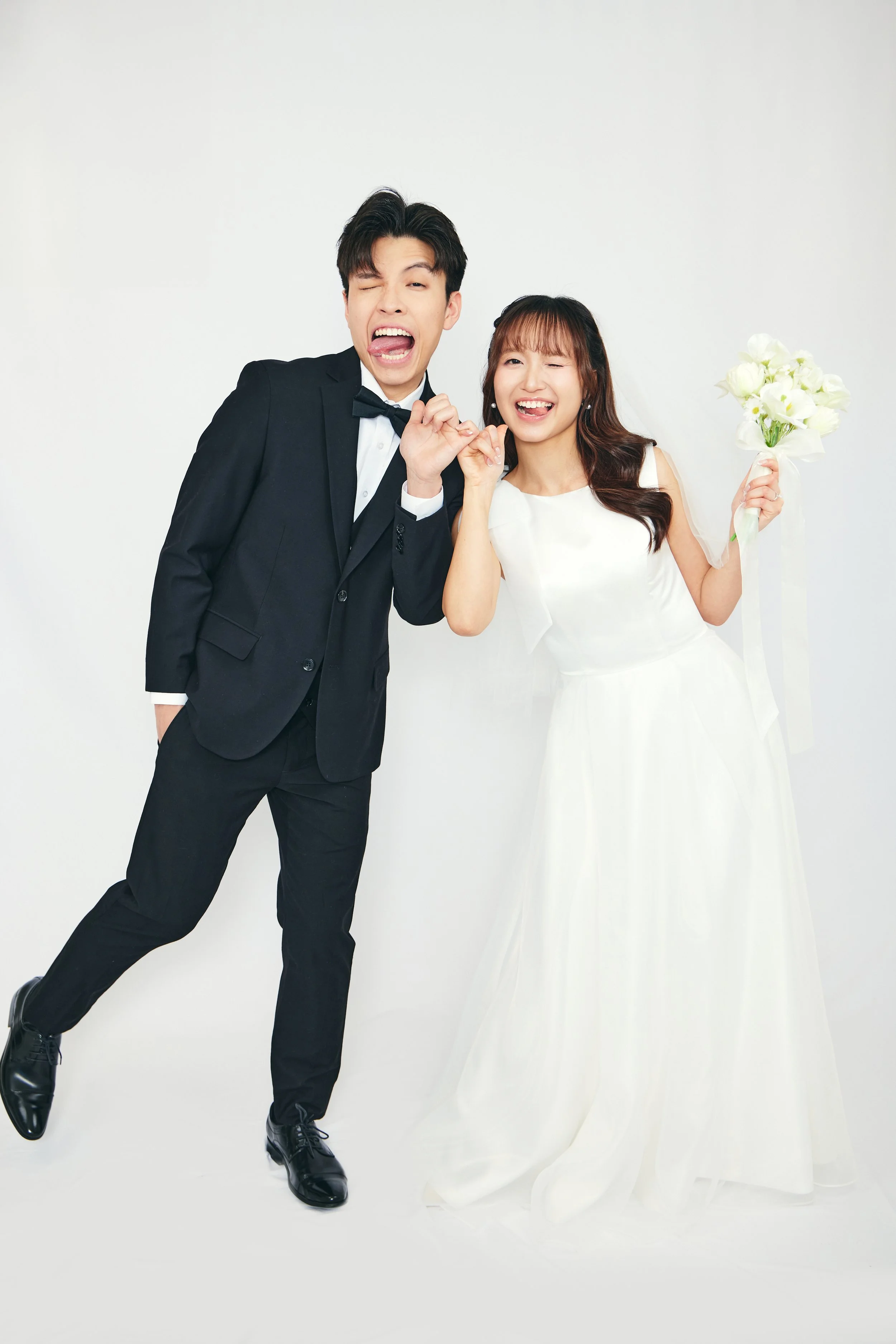 A happy bride in a white wedding dress holding a bouquet, and a groom in a black tuxedo making a playful pose against a plain white background.