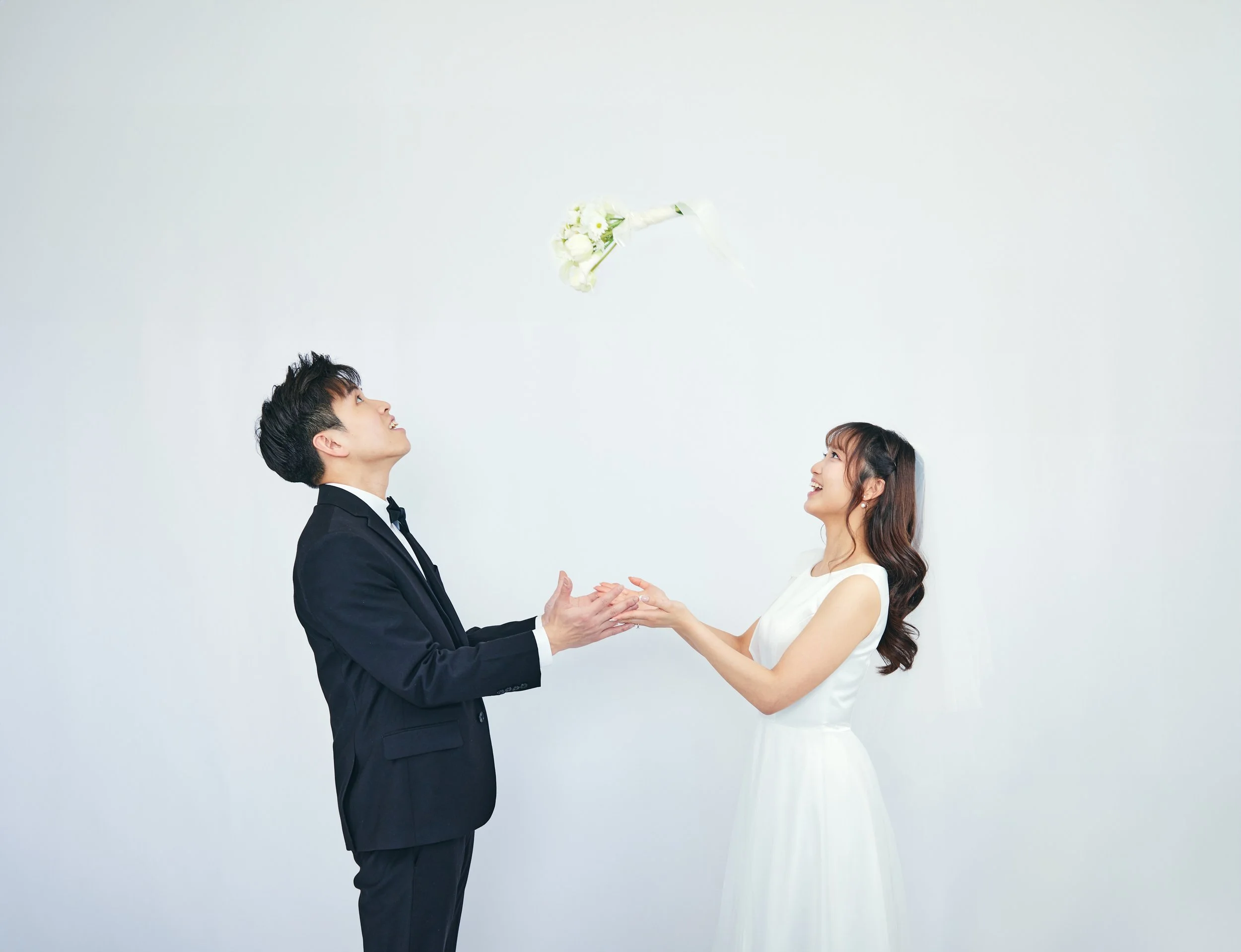 A bride and groom in wedding attire with the bride in a white dress and the groom in a black tuxedo, standing against a plain white background, as a bouquet of white flowers is tossed into the air between them.