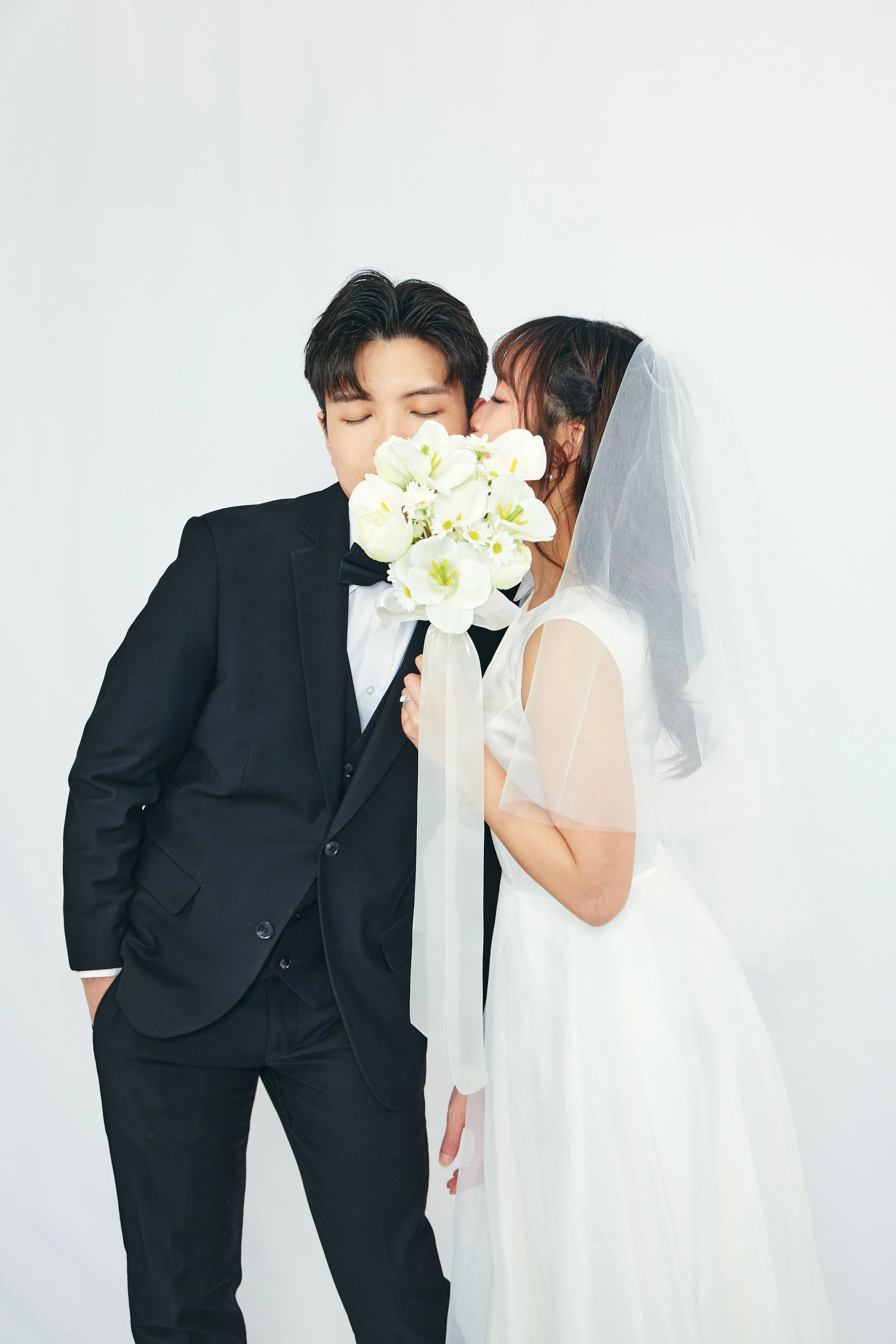 A bride and groom on their wedding day, with the bride kissing the groom's cheek while holding a bouquet of white flowers, against a plain white background.
