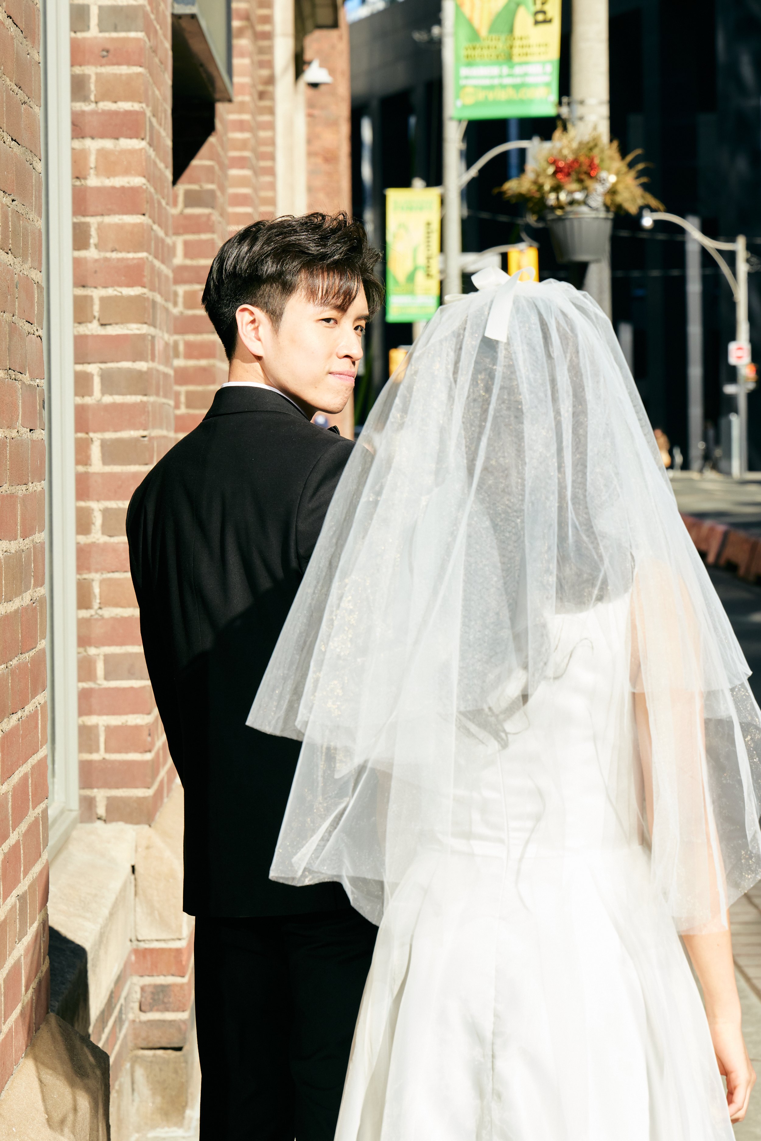 A man in a black suit looking at a woman in a wedding dress with a veil, standing on a city sidewalk near a brick building.