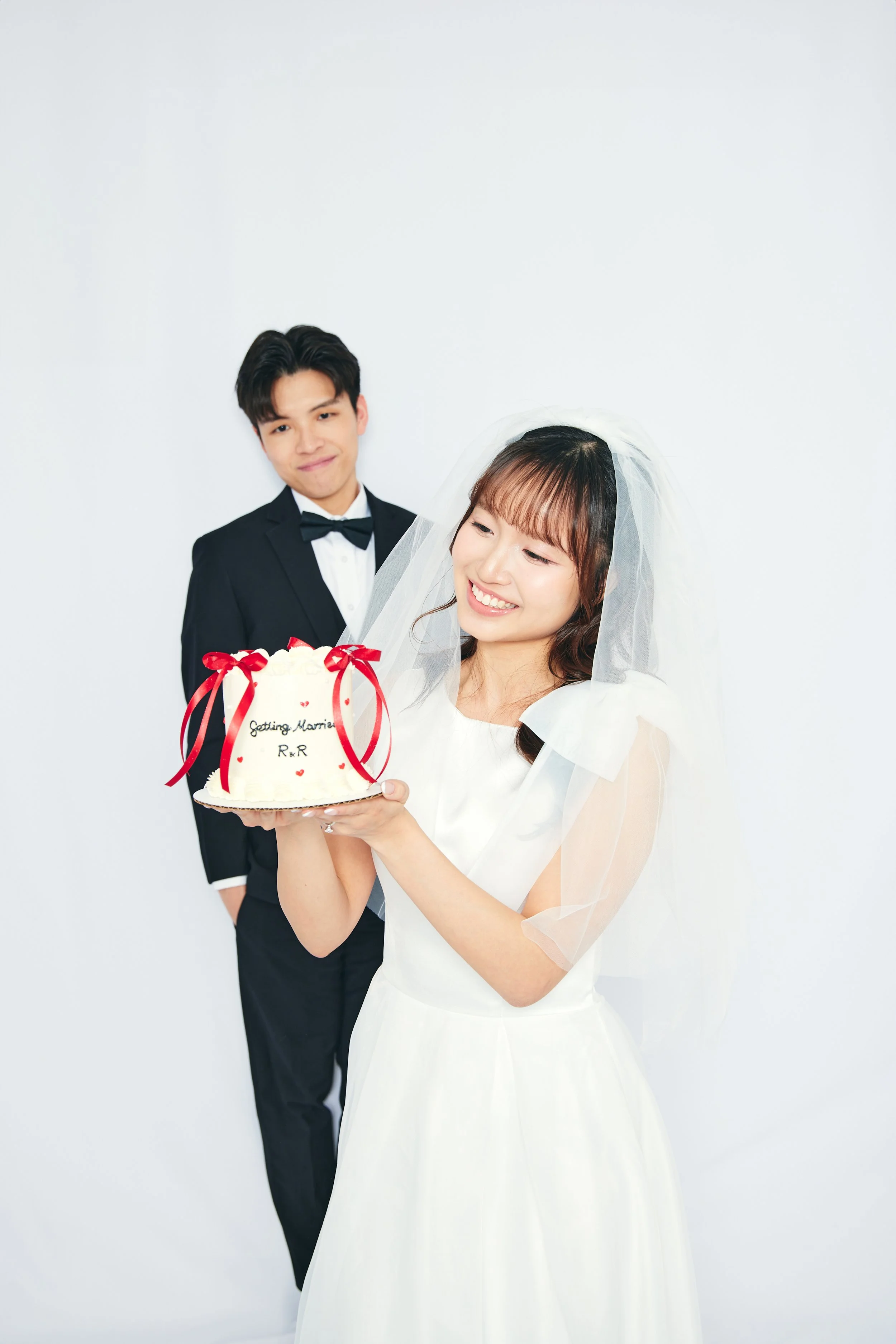 A bride in a white wedding dress and veil holding a wedding cake with red ribbons, while a groom in a tuxedo looks on, against a plain white background.