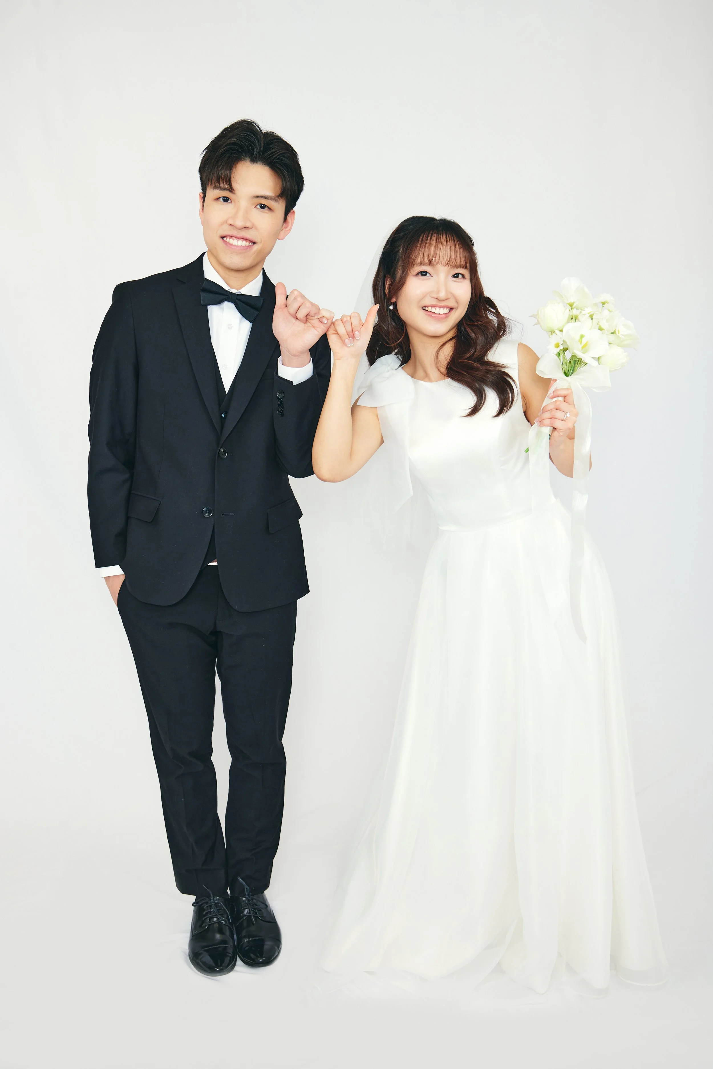 A newlywed couple in wedding attire posing together, with the groom in a black tuxedo and the bride in a white wedding dress holding a bouquet of white flowers.