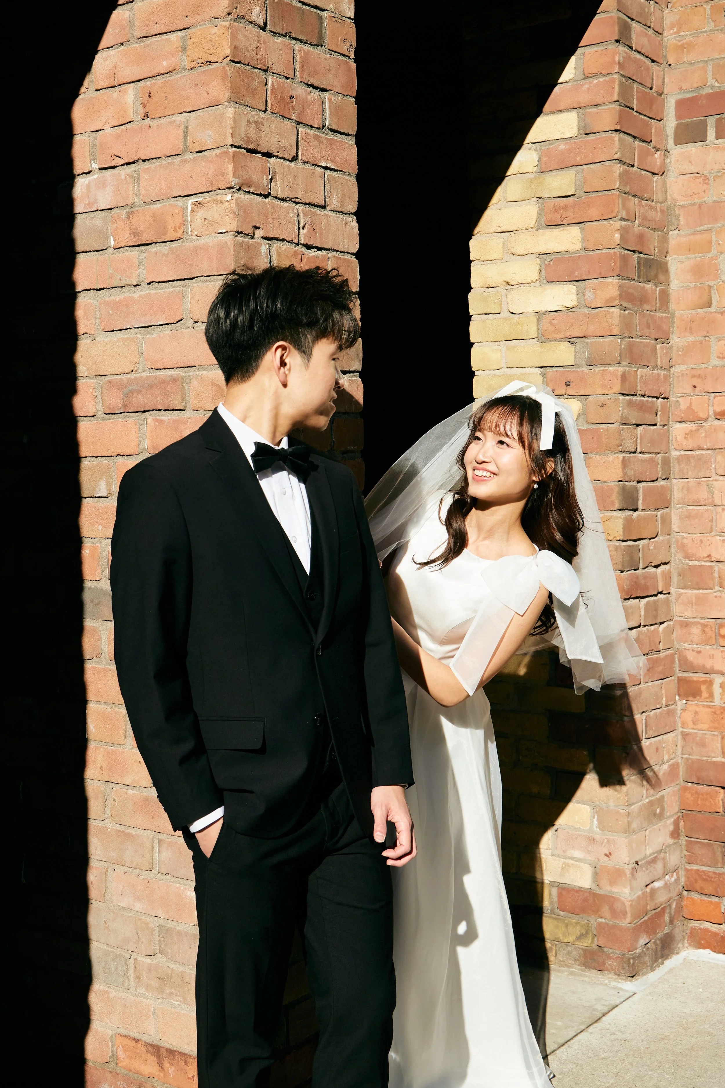 A bride and groom standing near a brick wall, looking at each other and smiling on their wedding day.