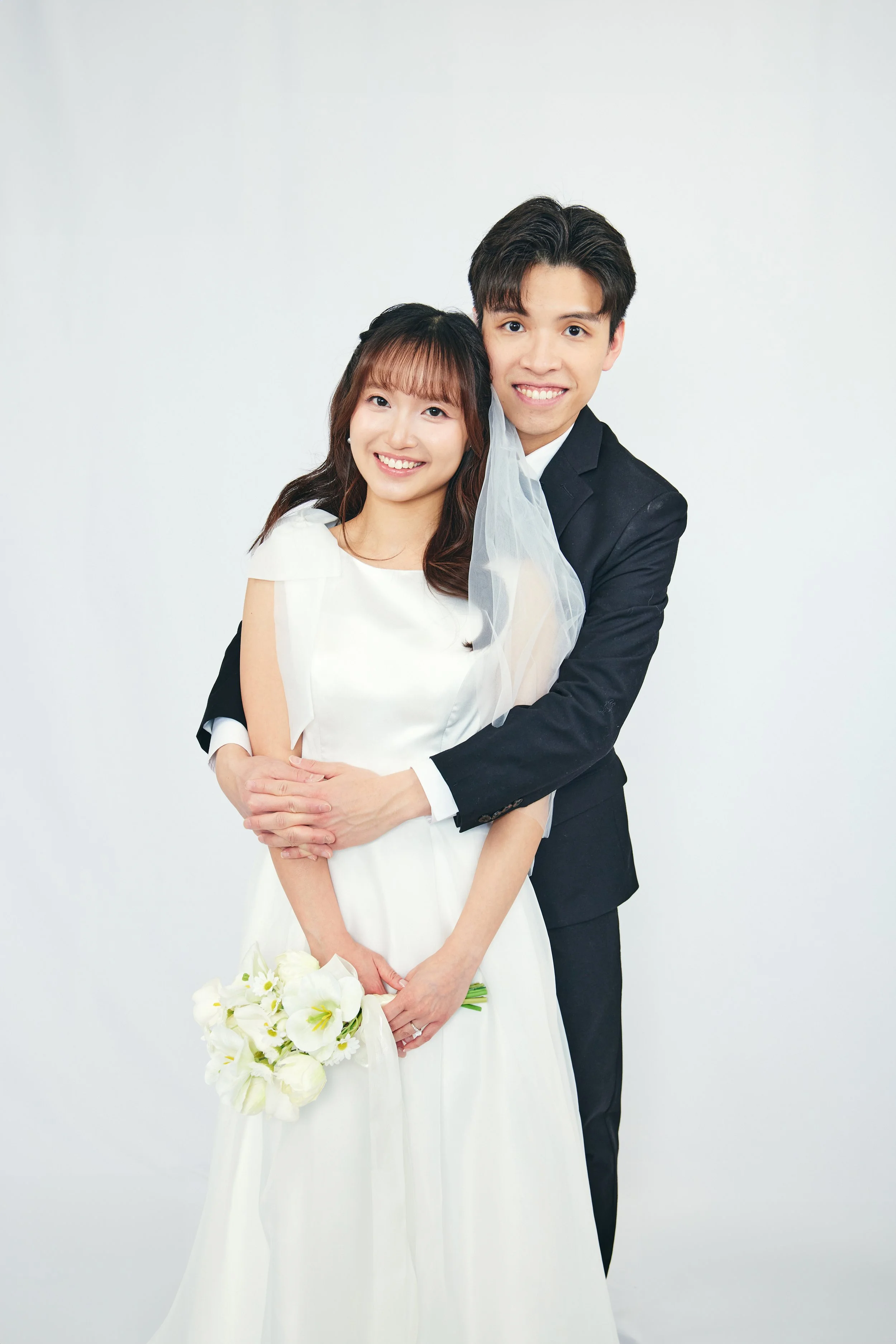 A smiling couple in wedding attire standing against a plain white background, embracing each other. The bride is holding a white bouquet and wearing a white dress with a veil, while the groom is dressed in a black suit.