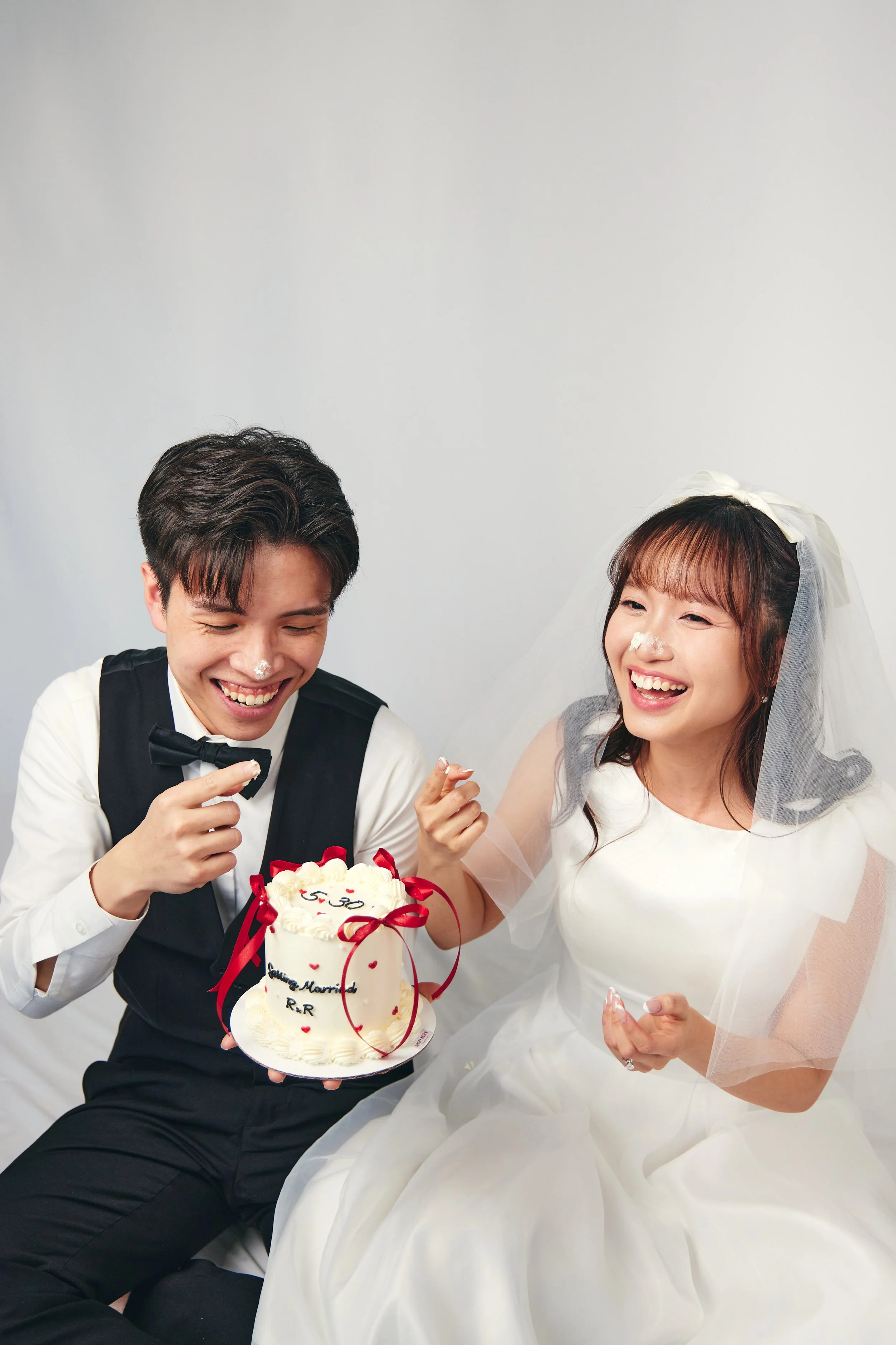 A happy couple in wedding attire celebrating with a cake, with frosting and decorations, and smeared frosting on their noses.