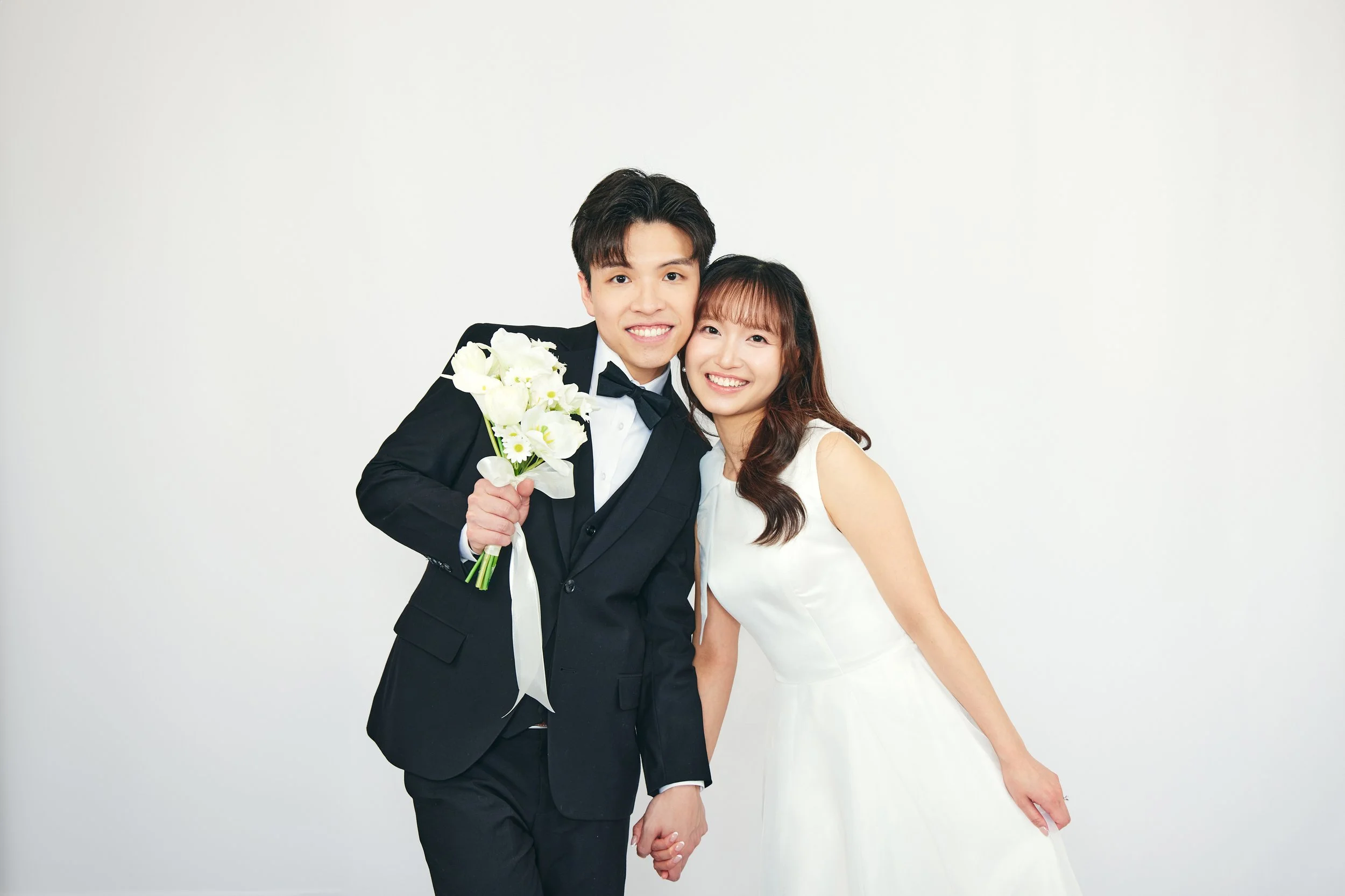 A young couple dressed in wedding attire, smiling and holding hands, with the groom holding a bouquet of white flowers.