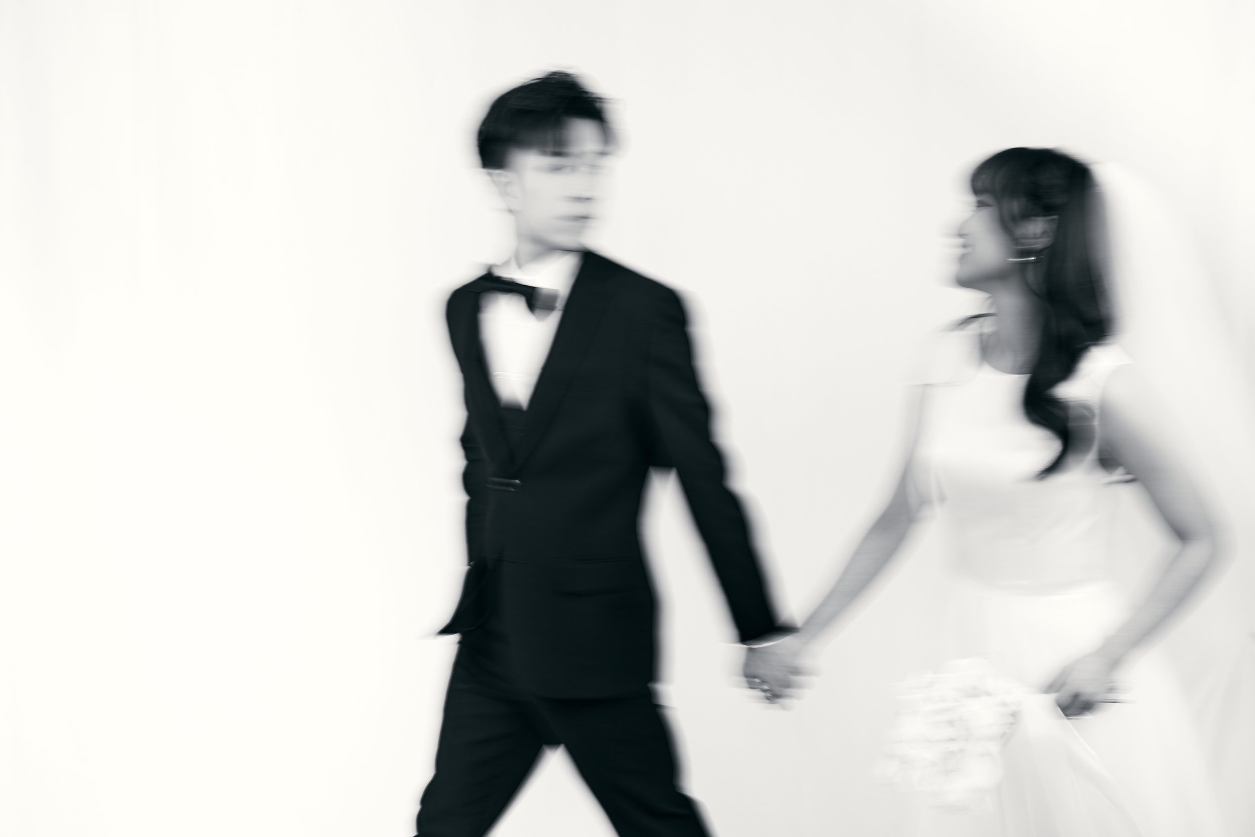 A blurry black and white photo of a man in a tuxedo holding hands with a woman in a white dress, both walking against a plain background.