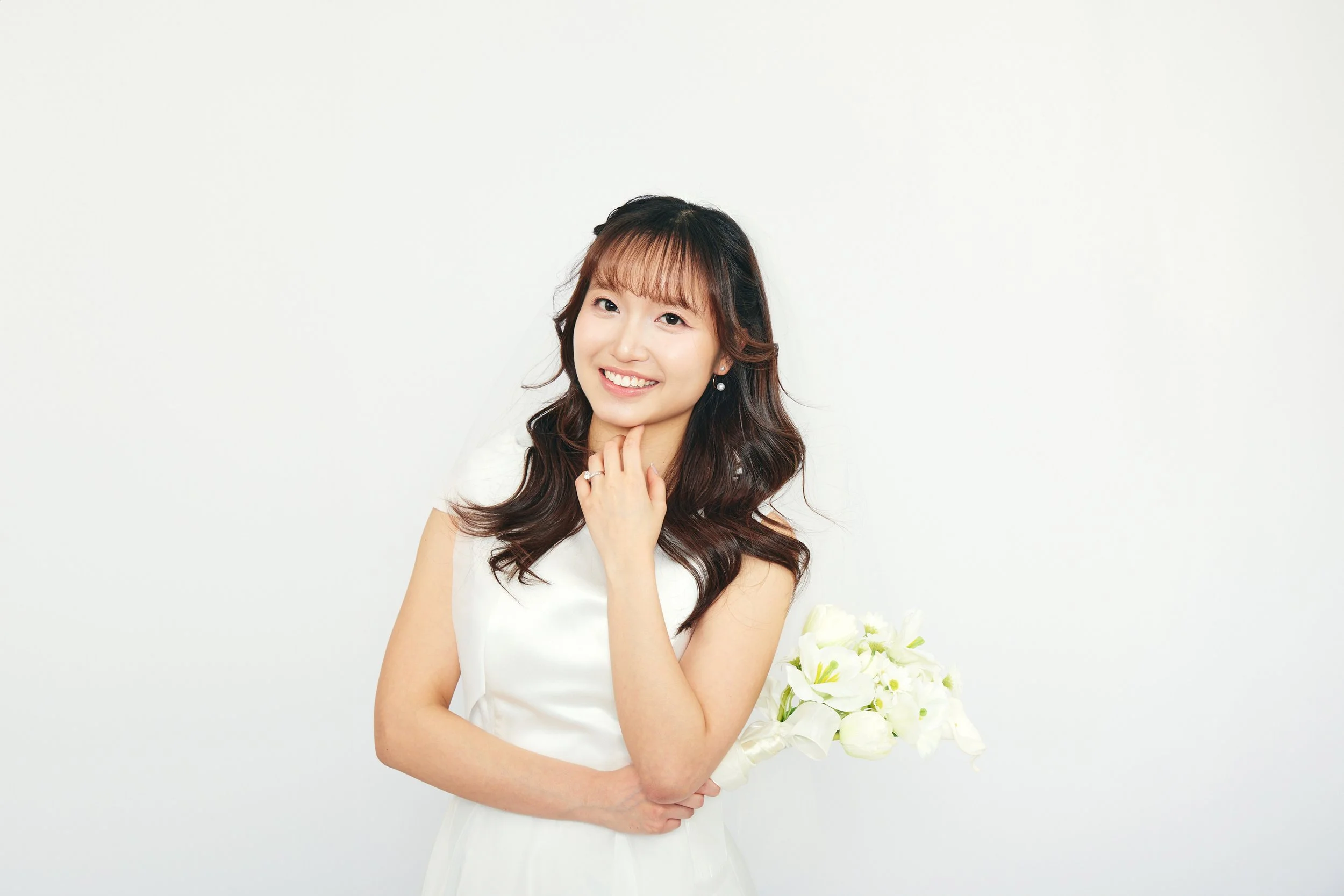 A young woman with long wavy brown hair wearing a white dress, holding a bouquet of white flowers, smiling against a plain white background.