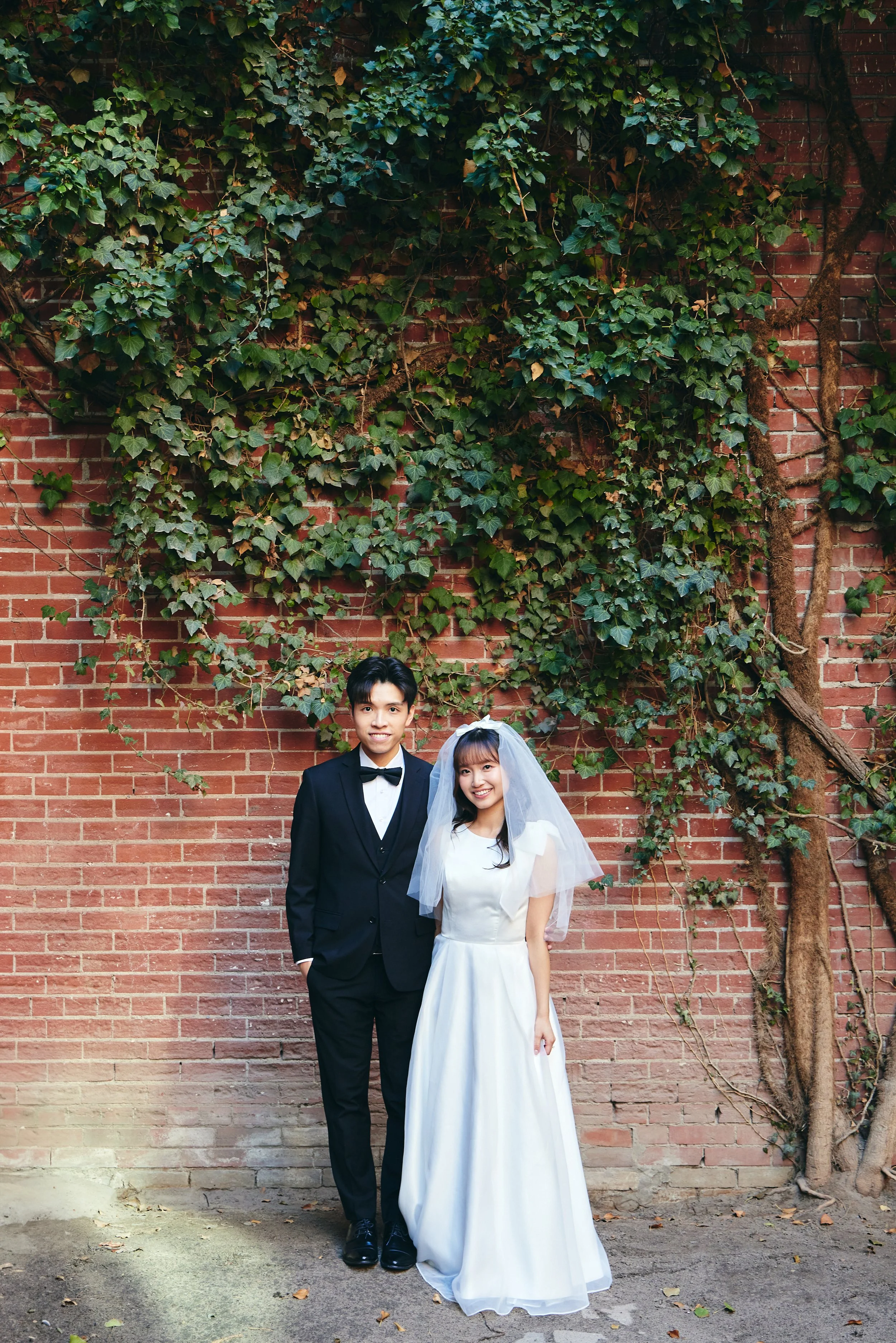 A wedding couple standing in front of a brick wall covered with green ivy, with the groom in a black tuxedo and the bride in a white wedding dress with a veil.