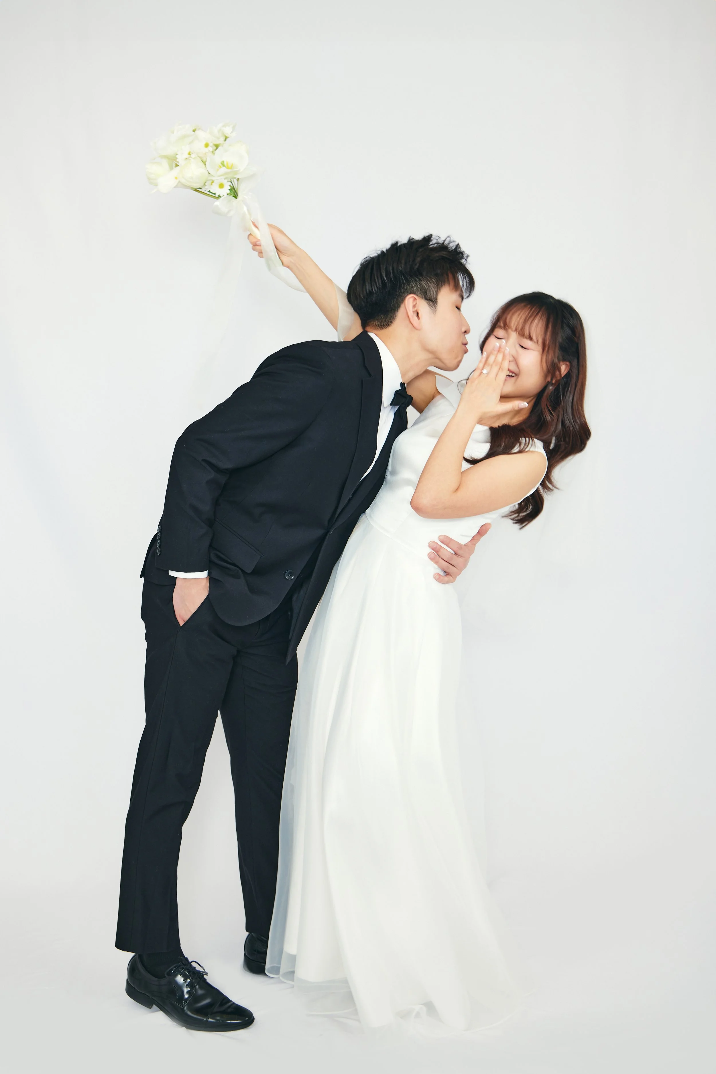 A bride and groom in wedding attire sharing a joyful moment, with the groom leaning in to kiss the bride on the cheek as she smiles and covers her mouth, against a plain white background. The groom holds a bouquet of white flowers behind his back.