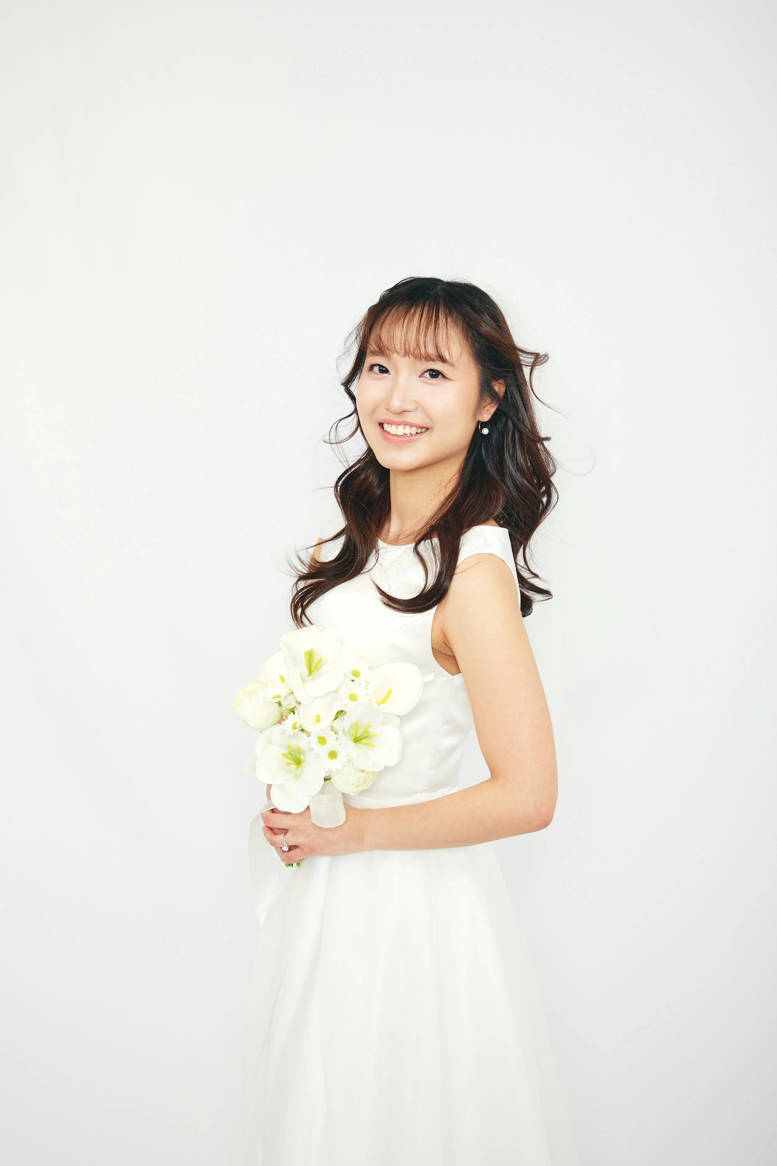 A young woman with long wavy dark hair in a white dress holding a bouquet of white flowers, smiling, standing against a plain white background.