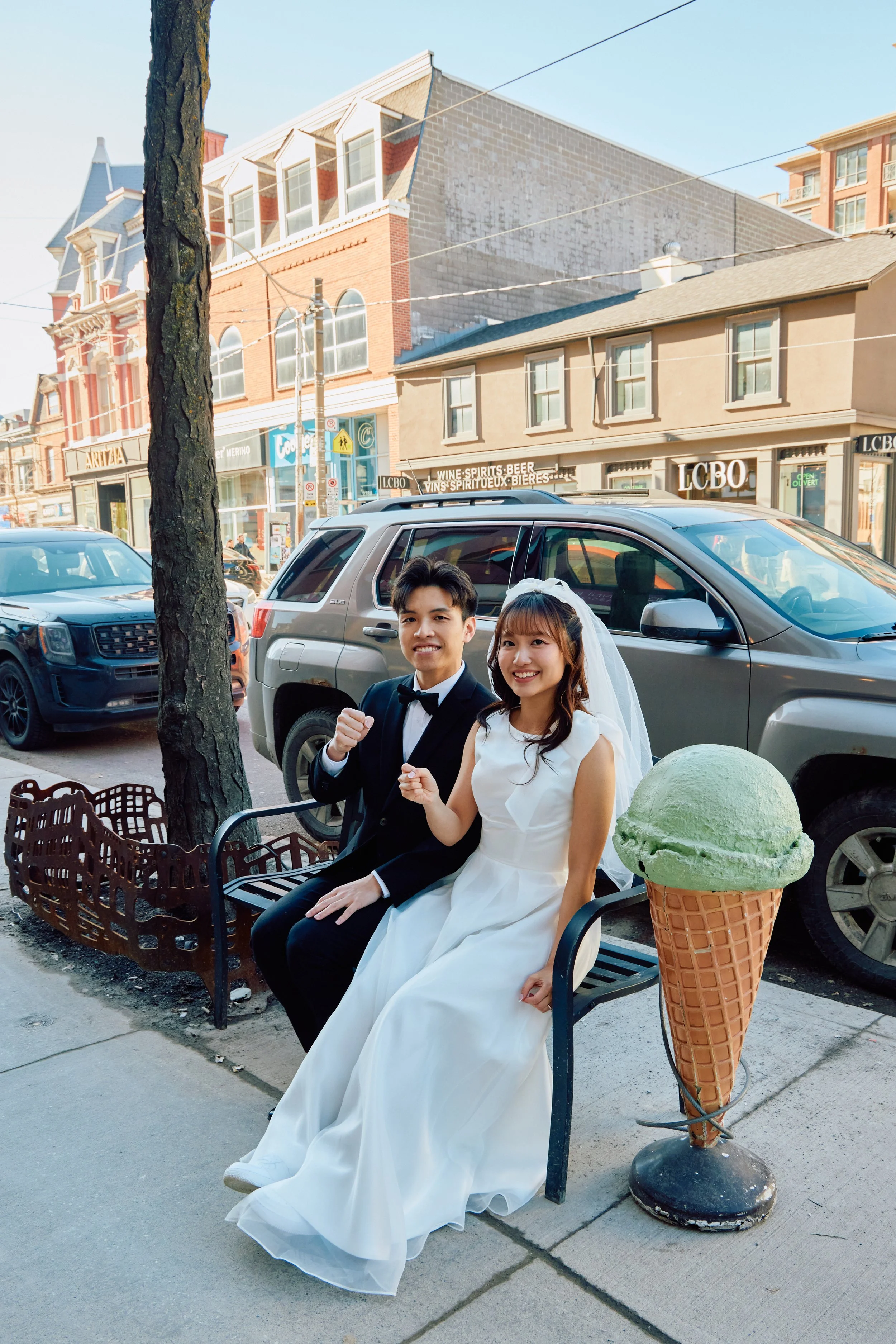 A couple dressed in wedding attire sitting on a bench outdoors in front of parked cars and buildings, with an oversized ice cream cone decoration nearby.