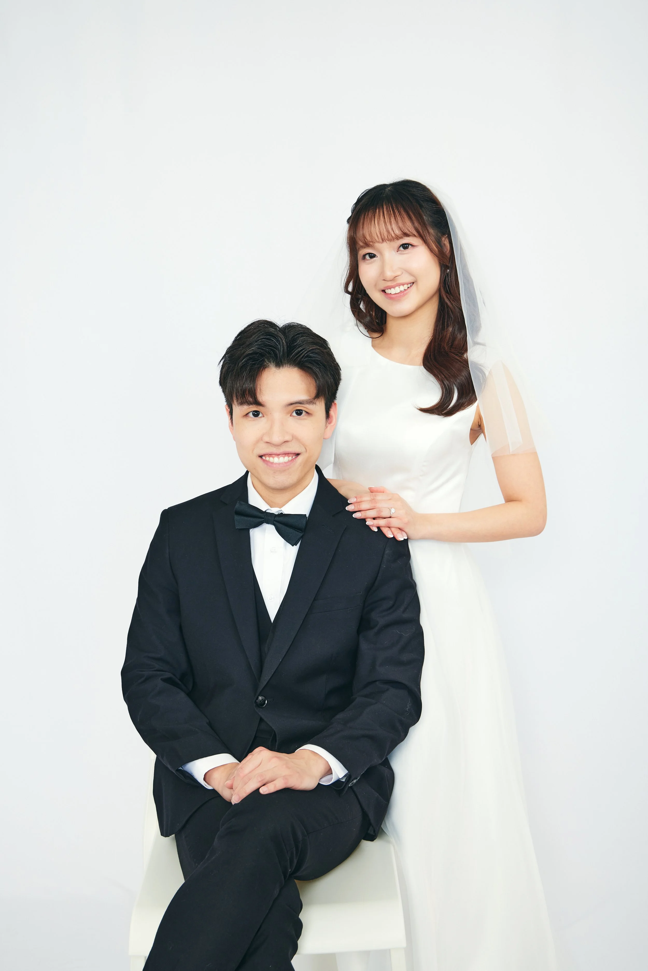 A young couple in wedding attire, the woman standing behind the seated man, both smiling, against a plain white background.
