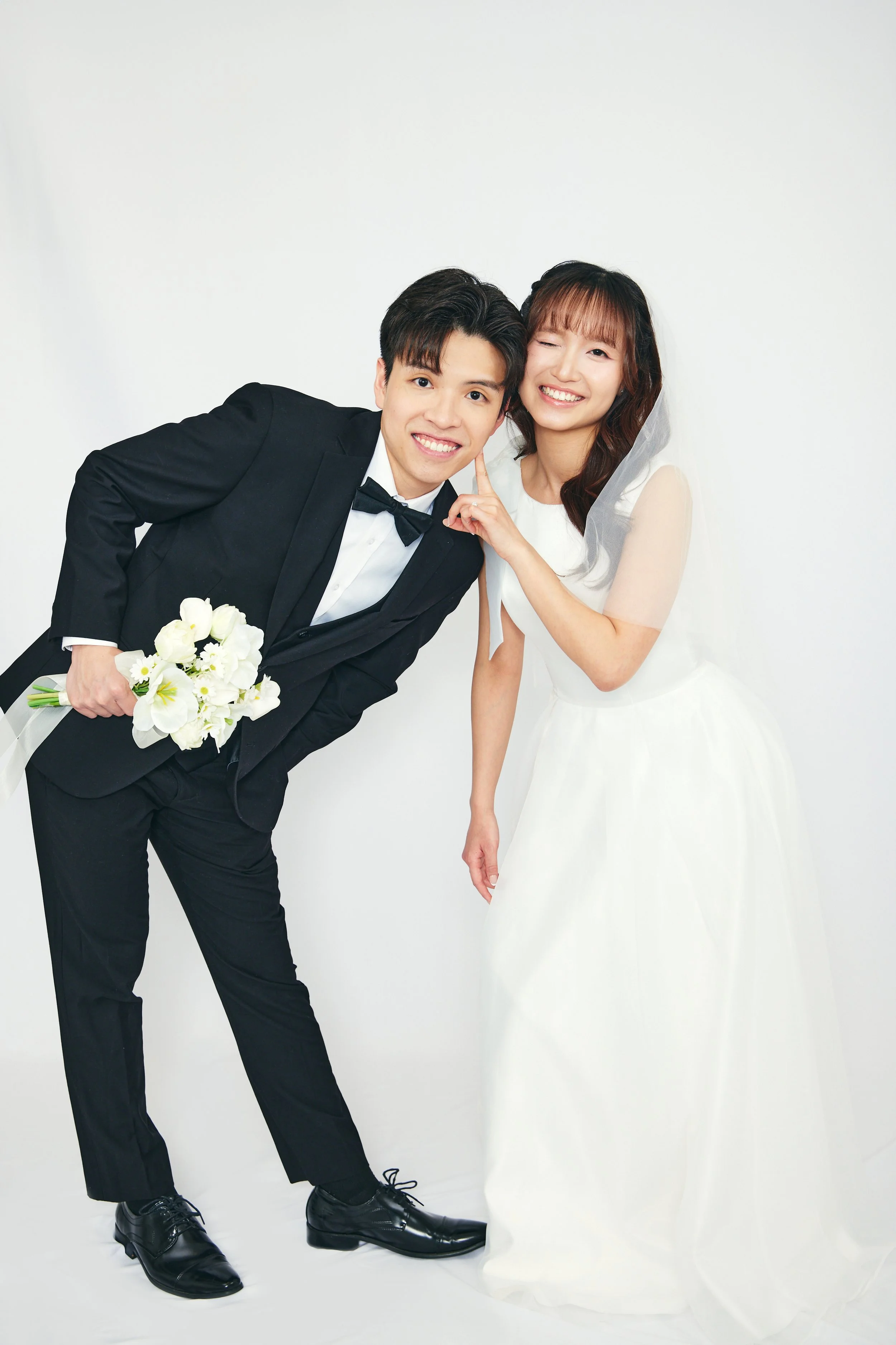 A smiling bride and groom posing together, the groom holding a bouquet of white flowers, standing against a plain white background.