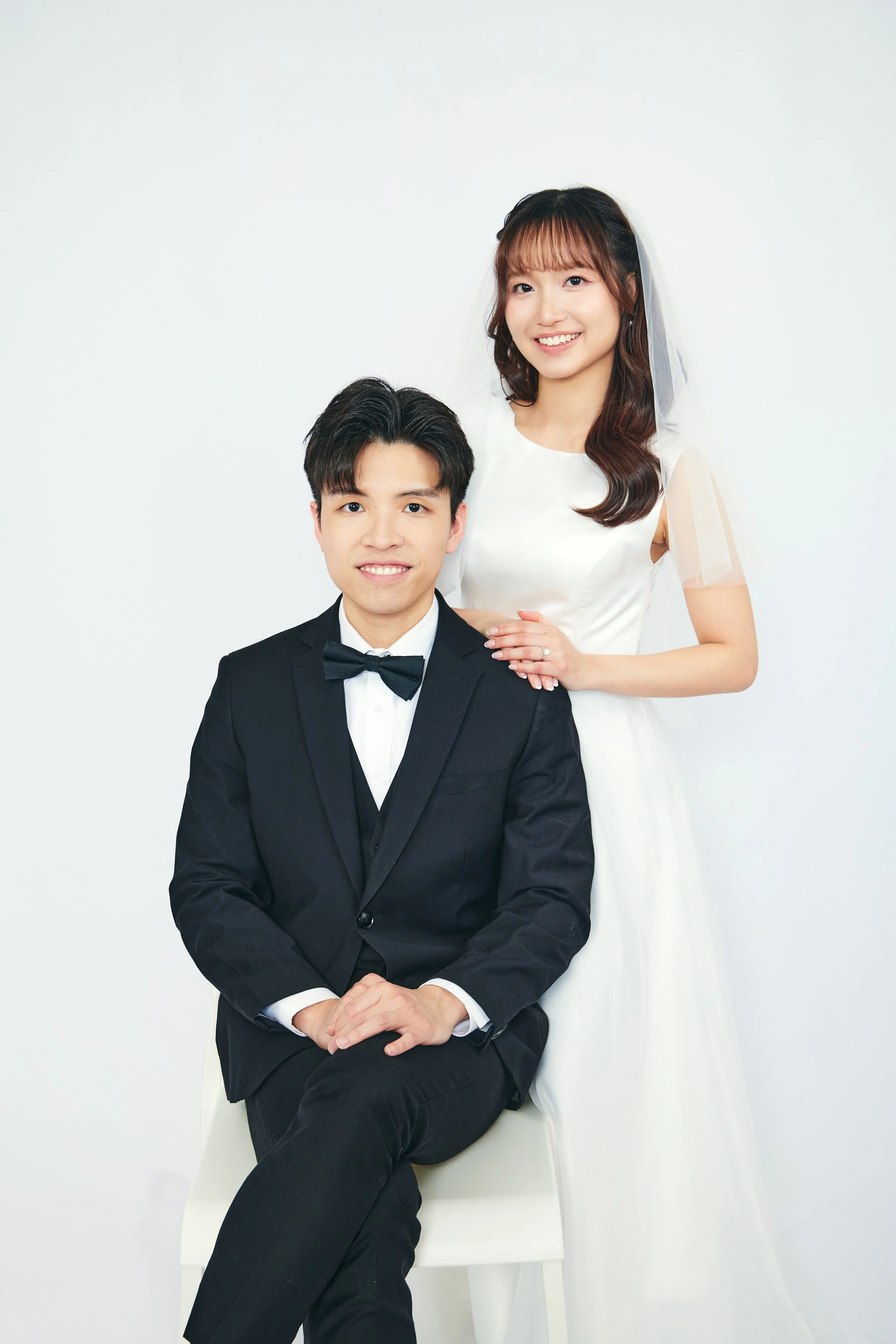 A bride and groom pose for a wedding photo against a plain white background. The groom, sitting on a white chair, wears a black tuxedo with a bow tie and white shirt, and the bride, standing behind him with a hand on his shoulder, wears a white weddi