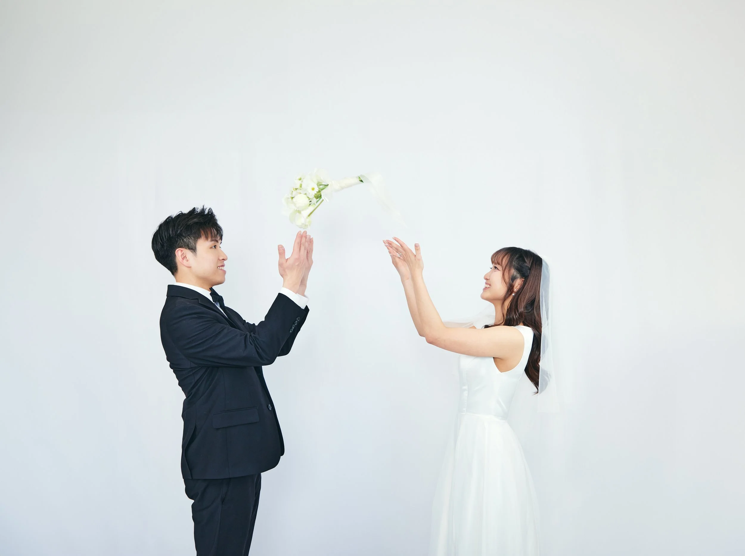 A bride in a white wedding dress and veil is smiling as she reaches out to catch a bouquet of white flowers, while a groom in a black suit and tie stands nearby with his hands raised, against a plain white background.