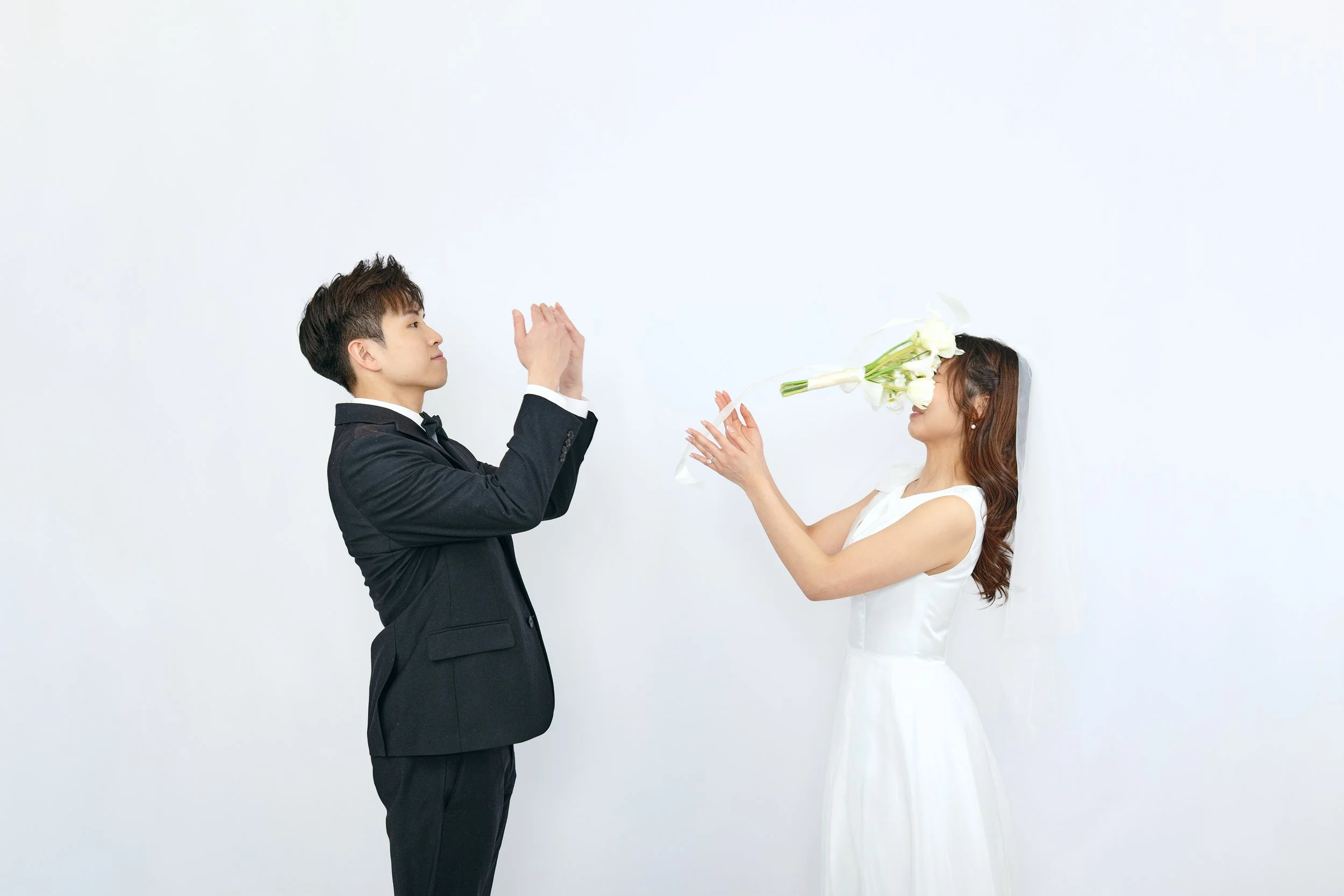 A groom in a black tuxedo appears to be clapping or praying while a bride in a white wedding dress holds a bouquet of white calla lilies over her face, standing against a plain white wall.