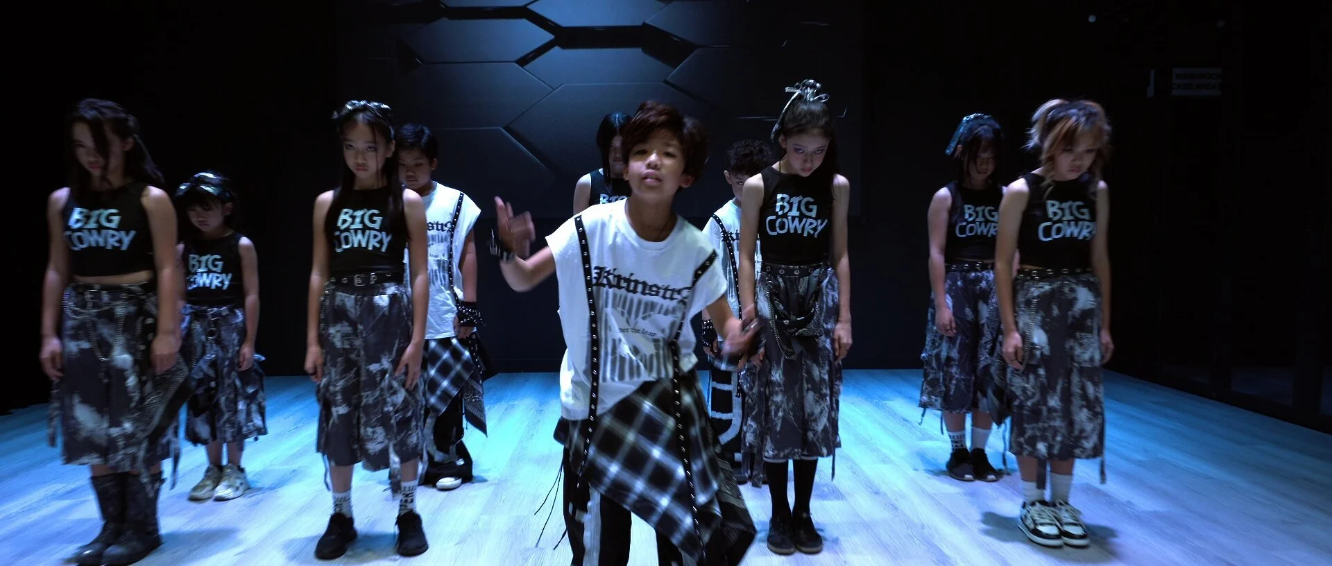 Frame from the One More Time music video: the dance team in matching black and grey outfits stands in a line on the studio stage under blue light.