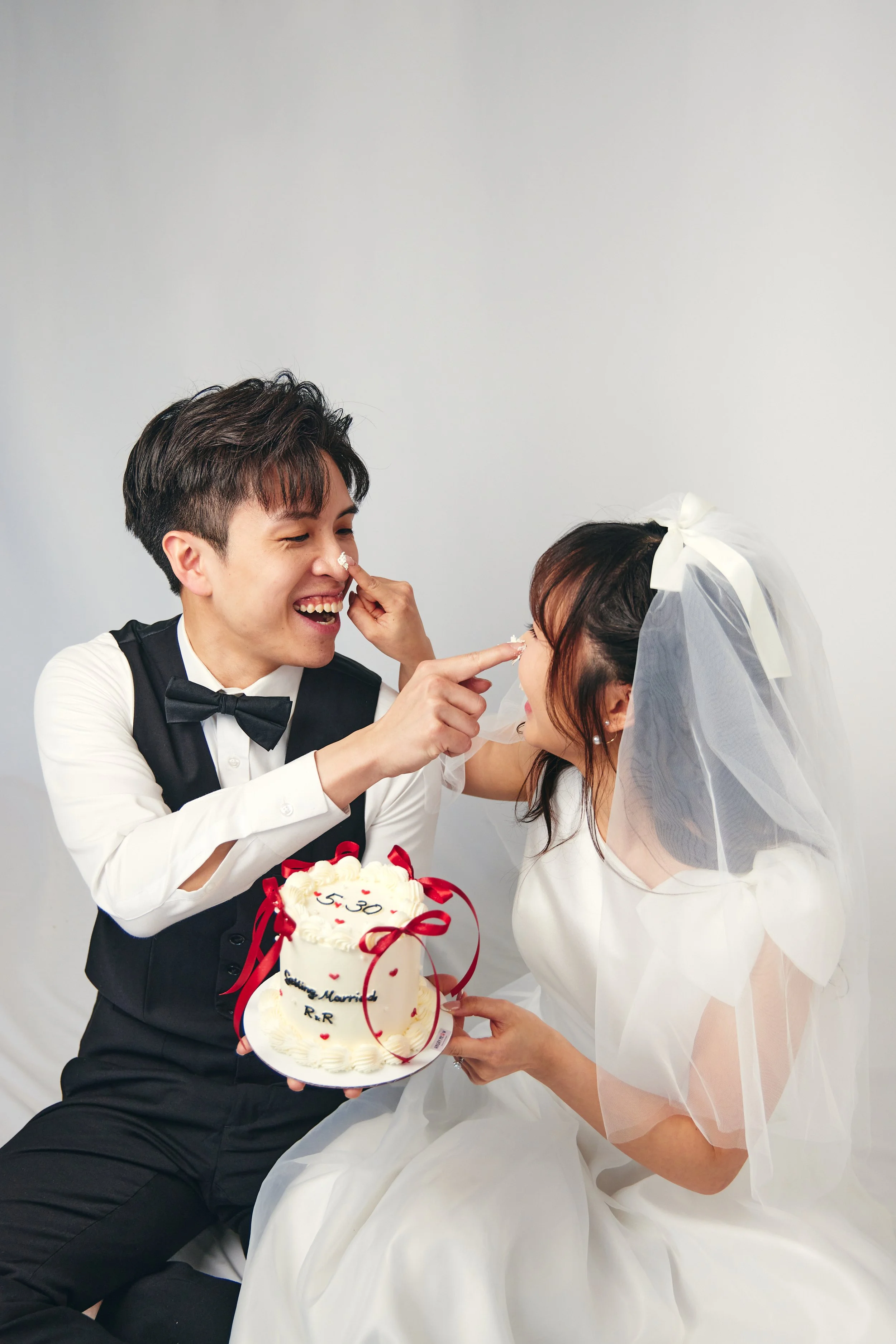 A newlywed couple celebrating their wedding, with the groom feeding the bride cake, both smiling, holding a wedding cake decorated with red ribbons, hearts, and writing.