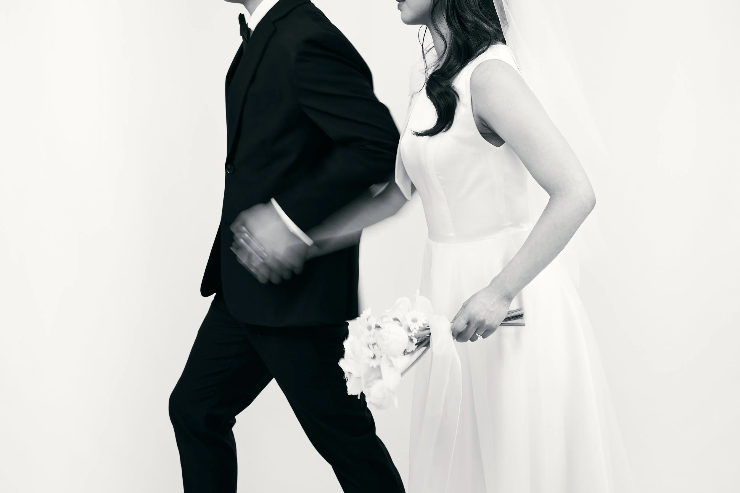 A black and white photo of a bride holding a bouquet and a groom with his back turned, both dressed in wedding attire.