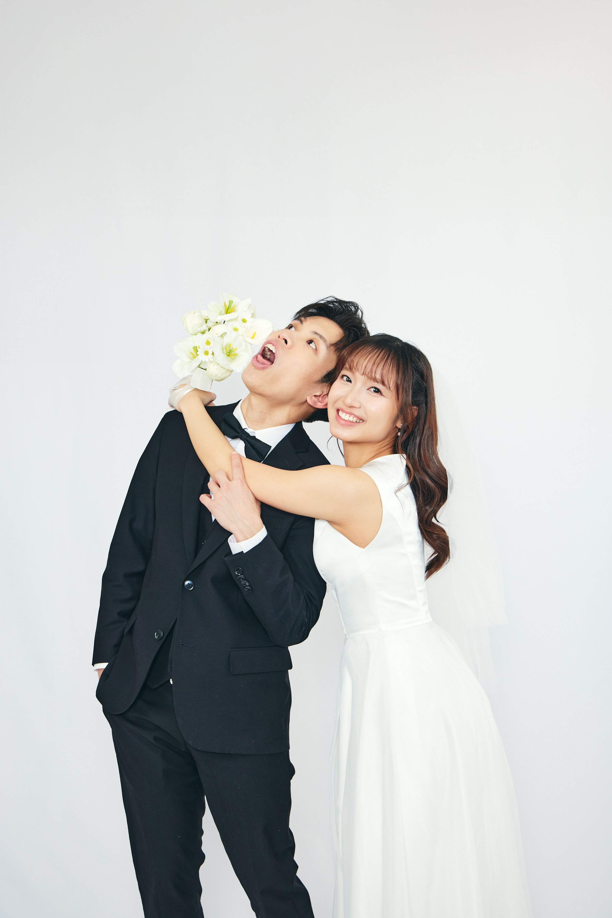 A newlywed couple in wedding attire playfully pose against a plain white background, with the bride hugging the groom from the side and holding a bouquet of white flowers near his face.