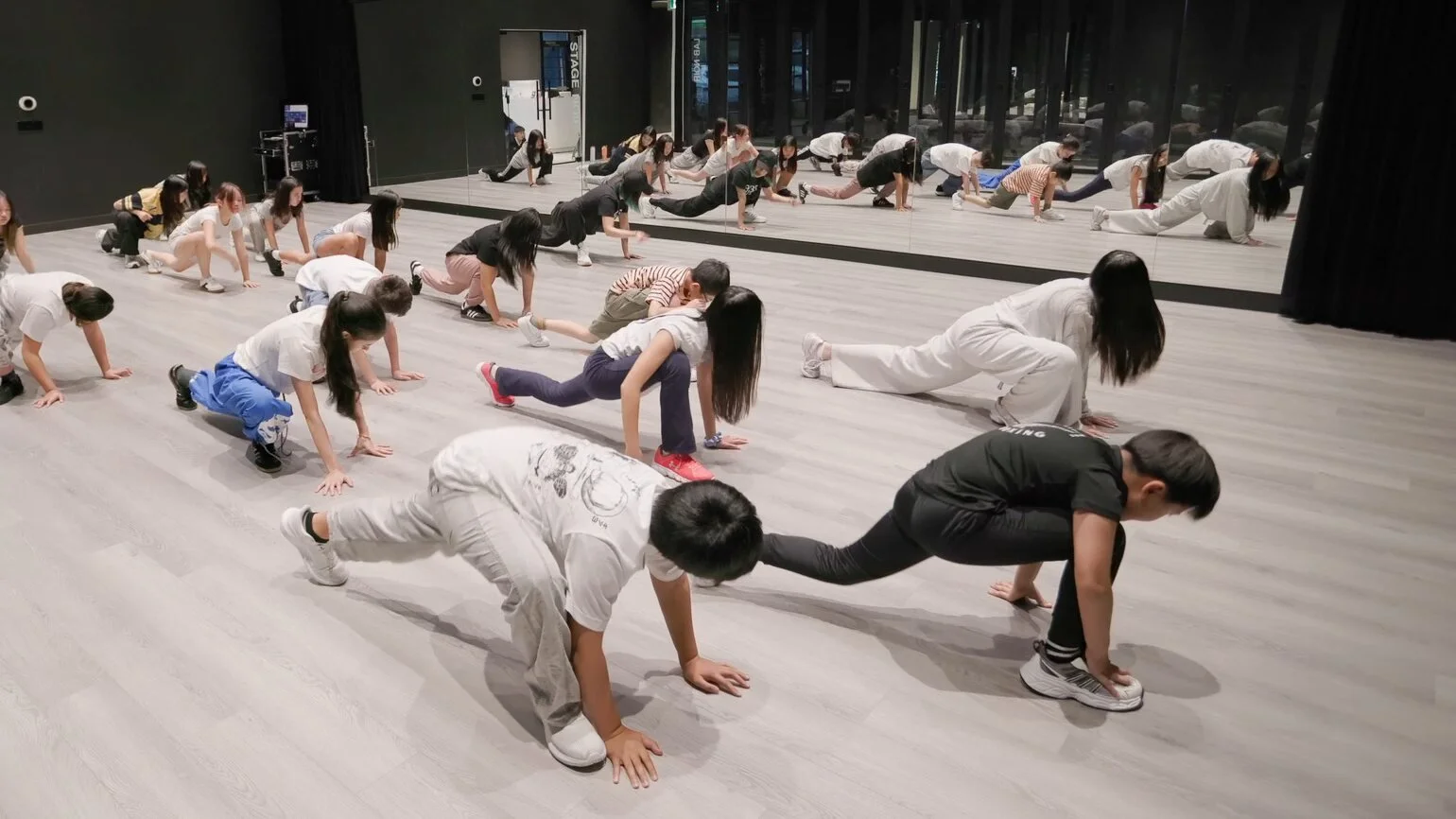 A wide shot of the full-year class warming up on the studio floor, low on their hands and feet in rows, with the mirror wall behind.
