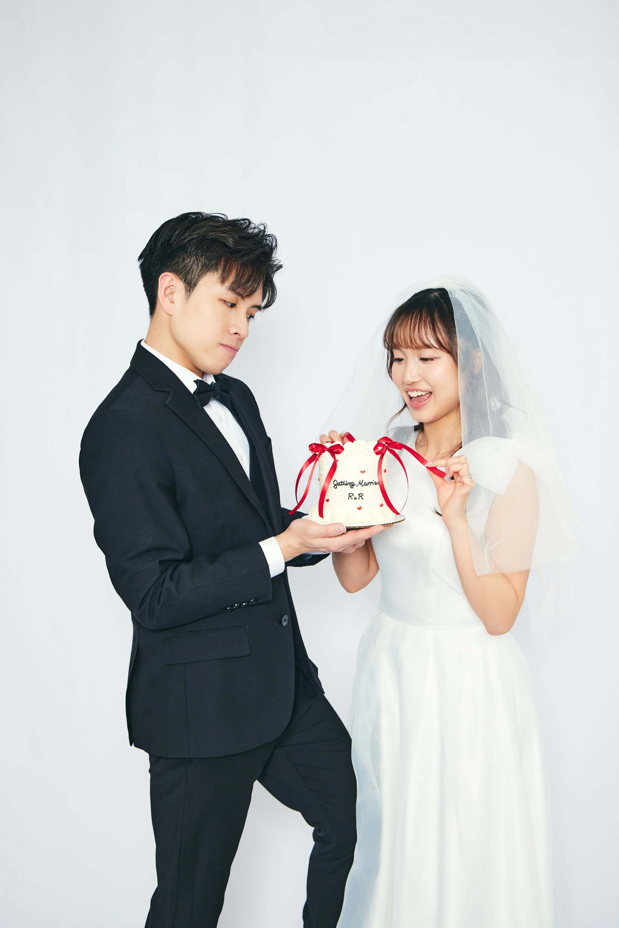 A bride and groom in wedding attire holding a small wedding cake decorated with red ribbons, smiling and looking at the cake.