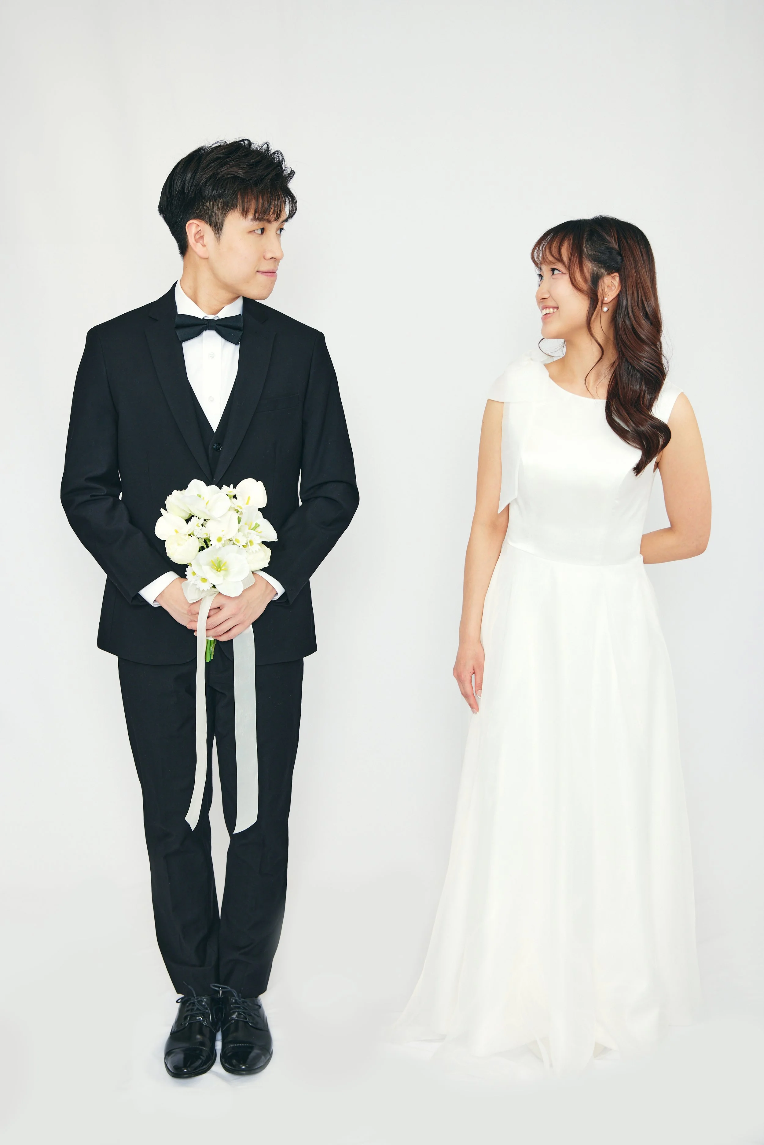 A groom in a black tuxedo holding a bouquet of white flowers looks at a bride in a white wedding dress who smiles back at him on a plain white background.