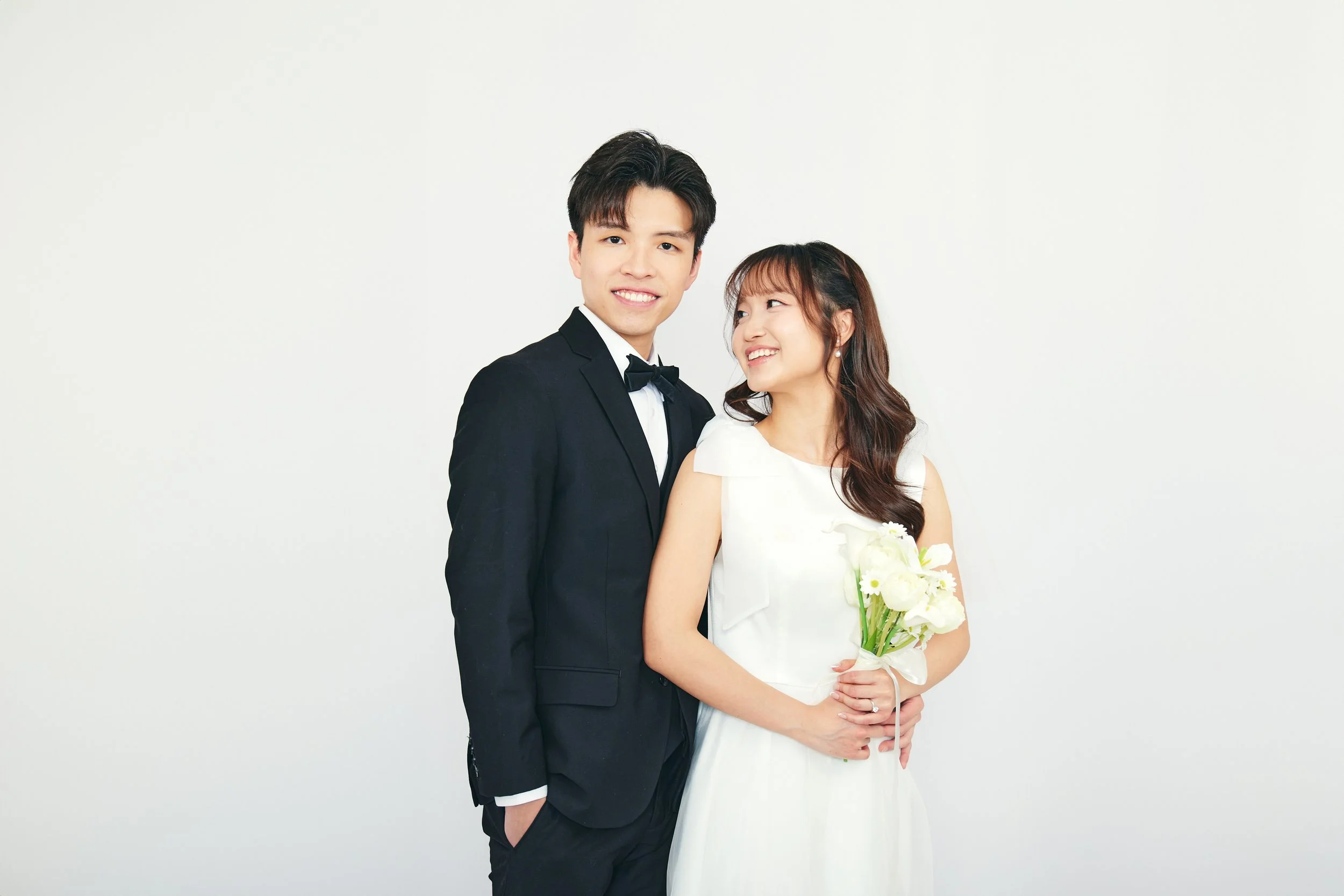 A smiling couple, dressed in formal wedding attire, standing against a plain white background, with the woman holding a bouquet of white flowers.