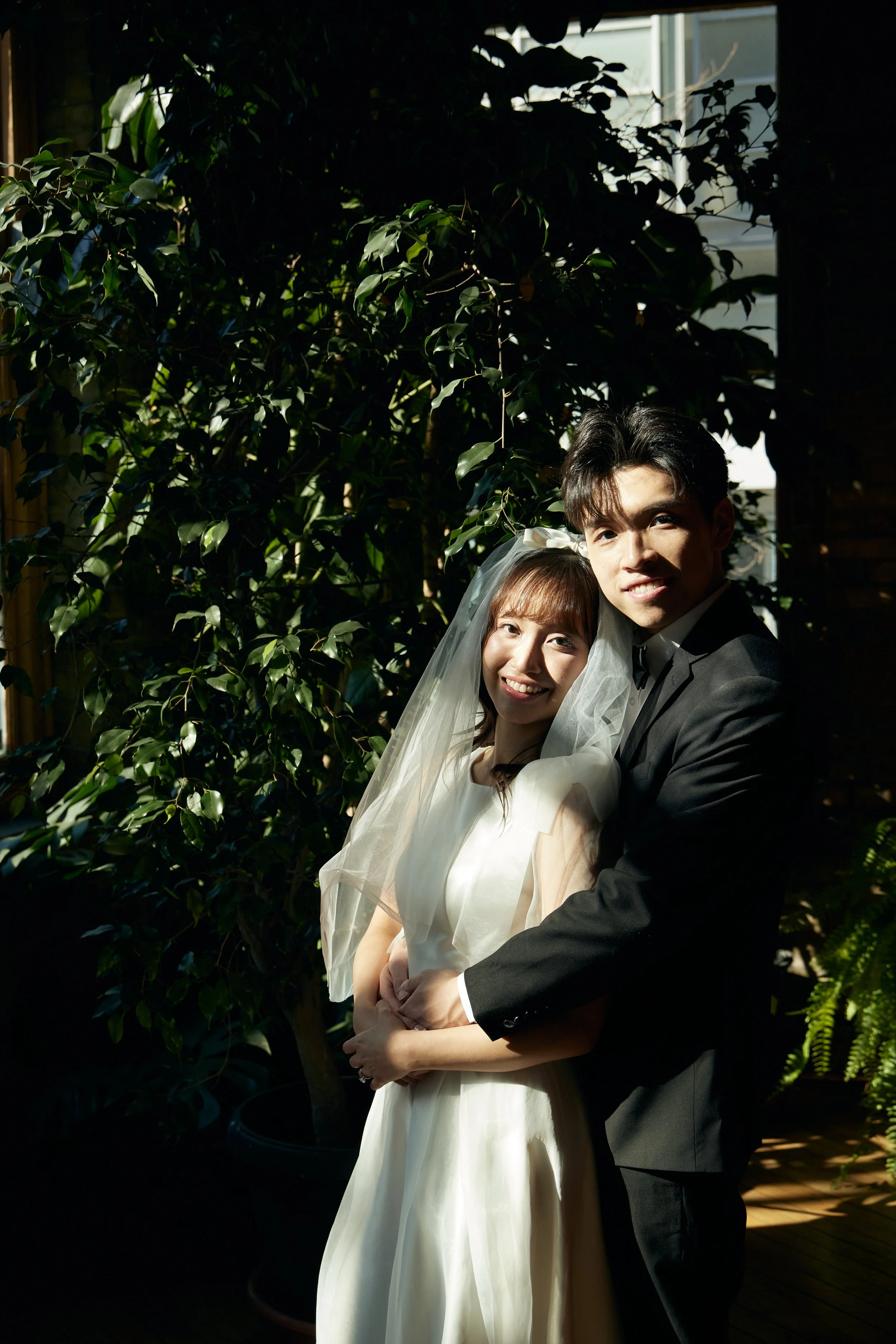 A bride and groom standing close together outside, embraced, smiling at the camera amid lush green foliage, with sunlight shining on their faces.