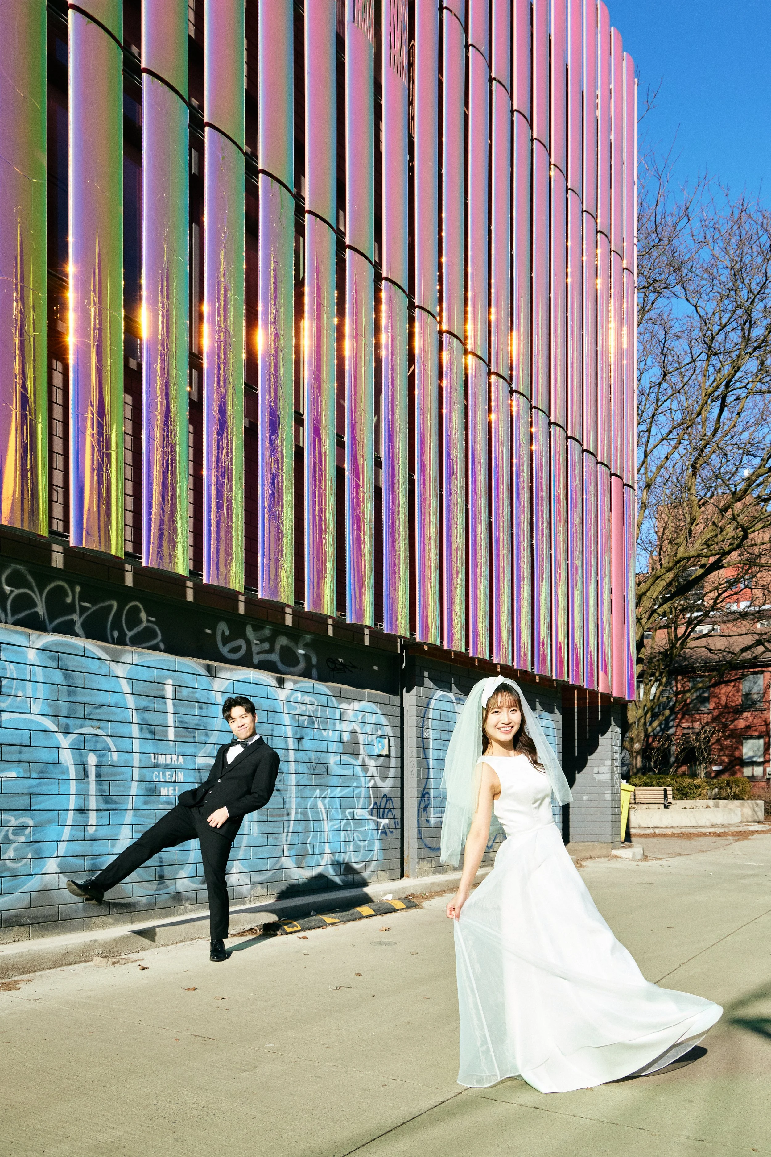 A couple in wedding attire posing outdoors in front of a graffiti-covered wall and an iridescent building under a clear blue sky. The bride is smiling in a white wedding dress and veil, while the groom is in a black tuxedo playfully kicking his leg.