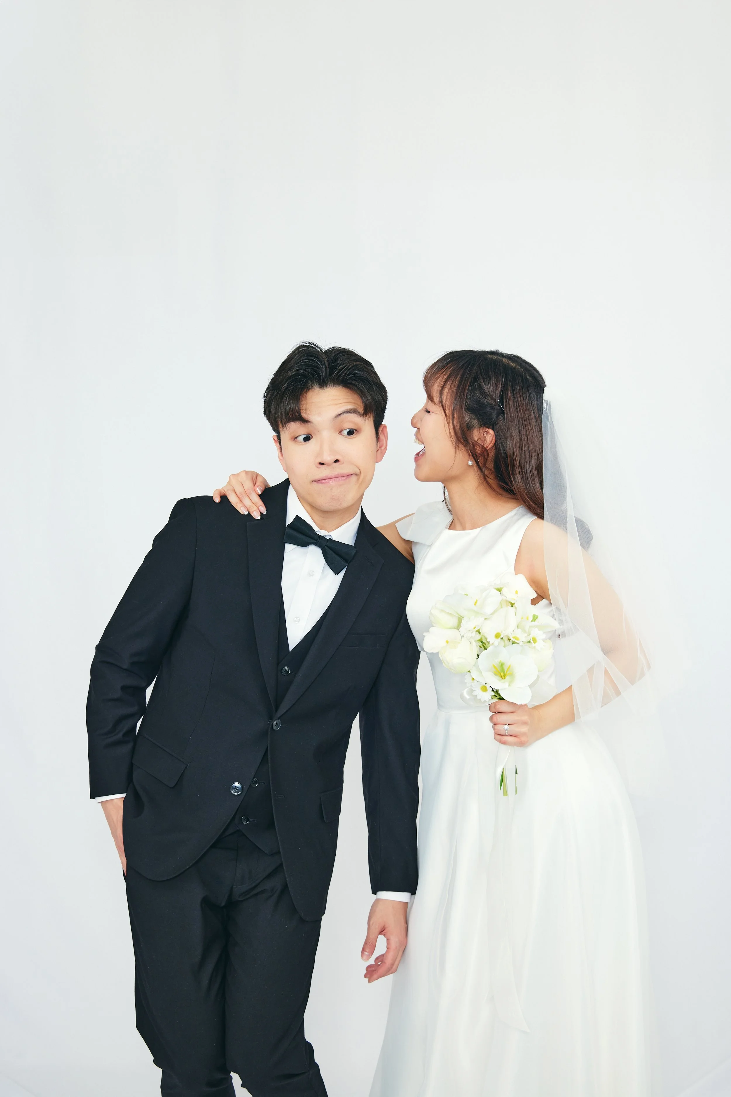 A bride and groom in wedding attire, with the bride holding a bouquet of white flowers, posing together against a plain white background.