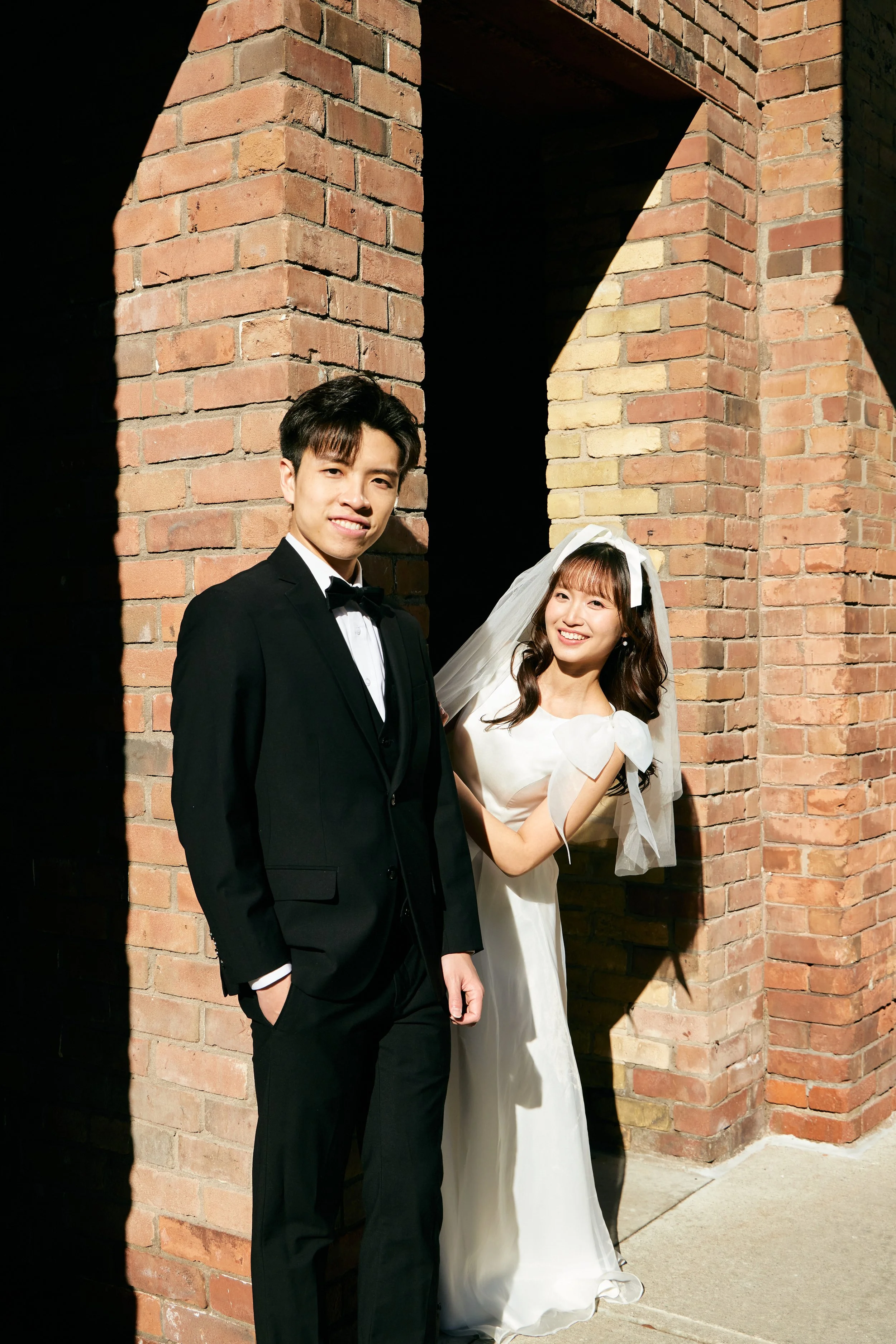 A smiling bride in a white wedding dress with bow details and a veil standing behind a groom in a black tuxedo with a bow tie, near a brick wall with sunlight and shadow.