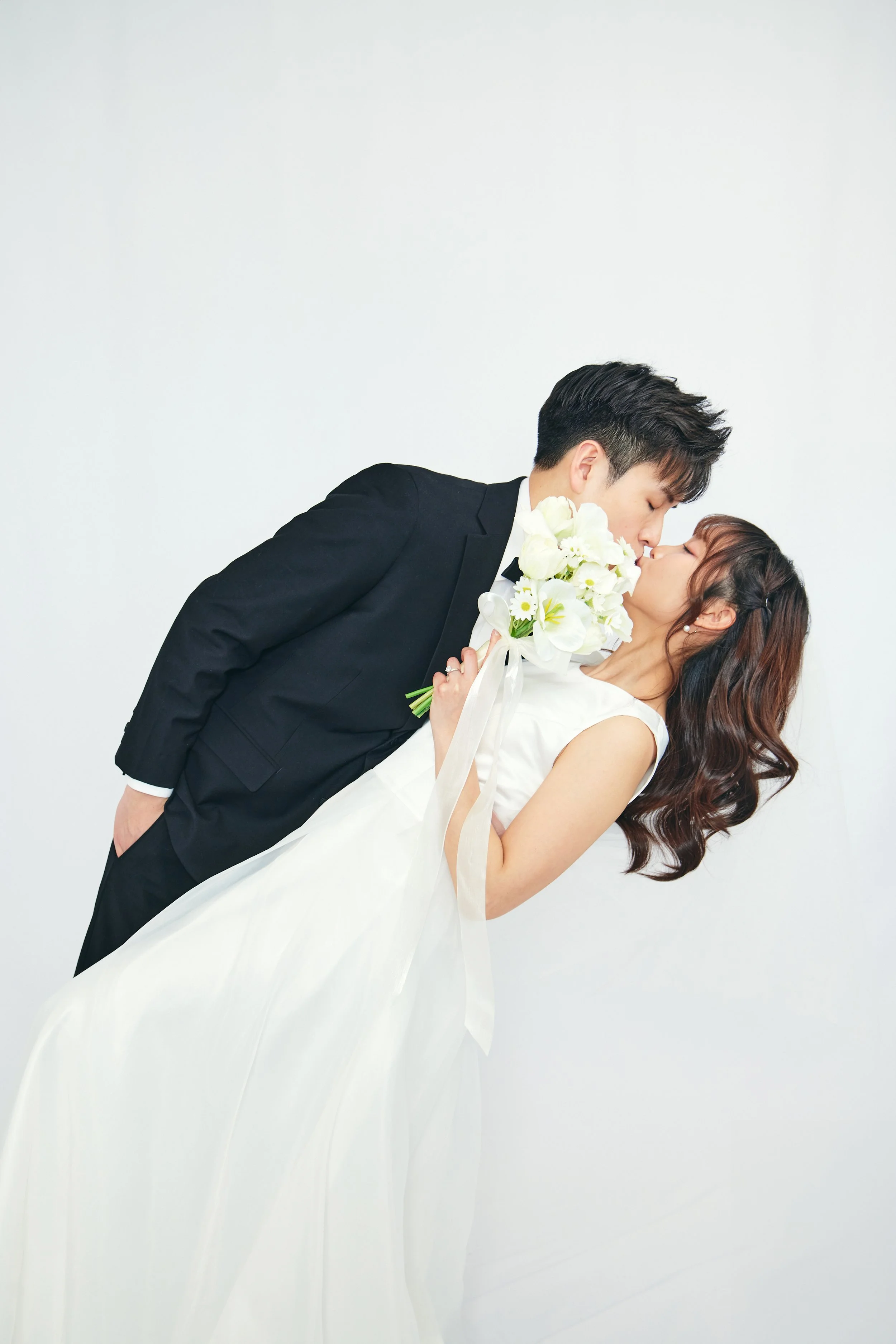 A bride and groom kiss, with the groom holding a bouquet of white flowers, posing against a plain white background.